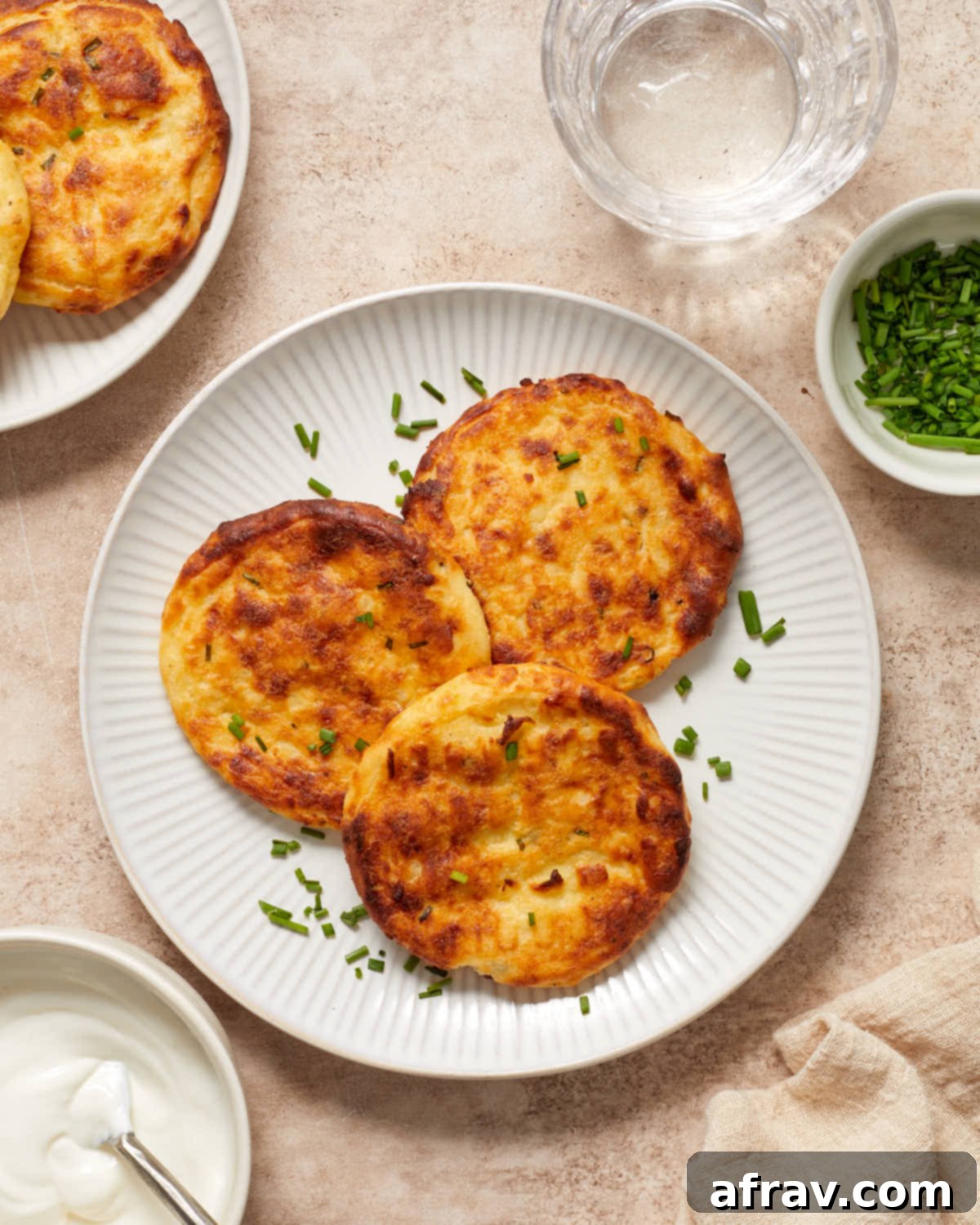 Three golden Air Fryer Potato Cakes on a plate, served with a side of sour cream, fresh chives, and a glass of water, highlighting a simple yet delicious meal.