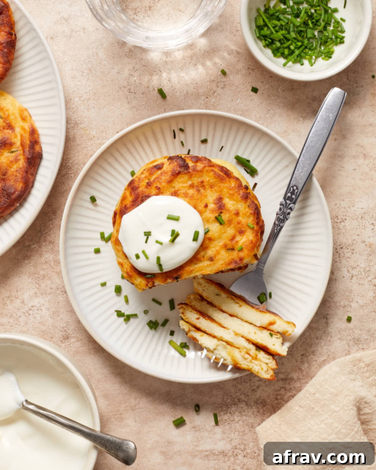 Overhead view of a stack of golden brown Air Fryer Potato Cakes on a white plate, elegantly topped with a dollop of sour cream and fresh chopped chives.