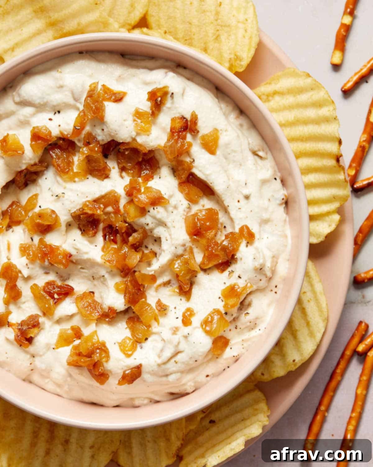 Overhead shot of a bowl of dairy-free onion dip surrounded by an assortment of potato chips and crunchy pretzels, ideal for sharing.