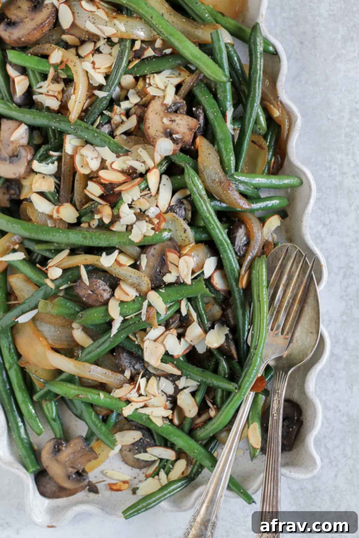 Green beans with sauteed onions and mushrooms sprinkled with sliced onions on a serving tray with a vintage fork and spoon laying on the tray.
