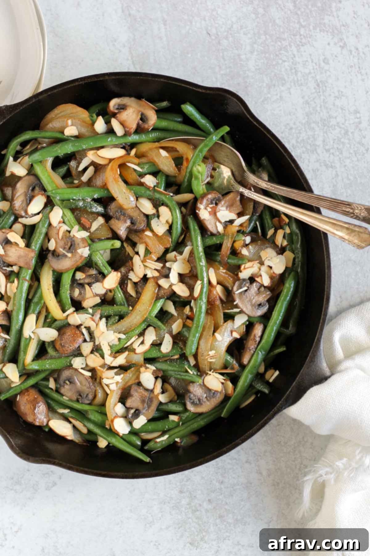 Savory Skillet Green Beans with Onions and Mushrooms 10 Green beans with mushrooms and onions topped with sliced almonds in a cast iron skillet with a vintage serving spoon and fork in the skillet.