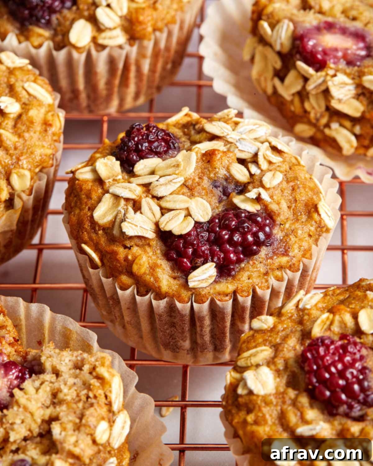 A close-up photograph of a single banana blackberry oatmeal muffin, showcasing its golden-brown top, visible oats, and soft texture, resting on a wire rack.