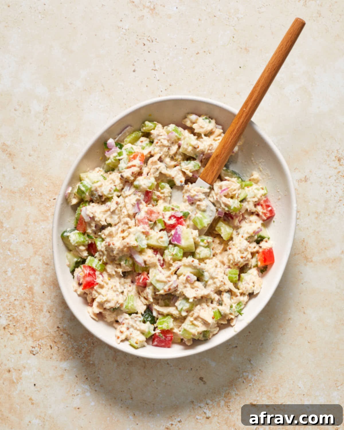 Freshly mixed tuna salad, vibrant with chopped red pepper and cucumber, in a white mixing bowl.