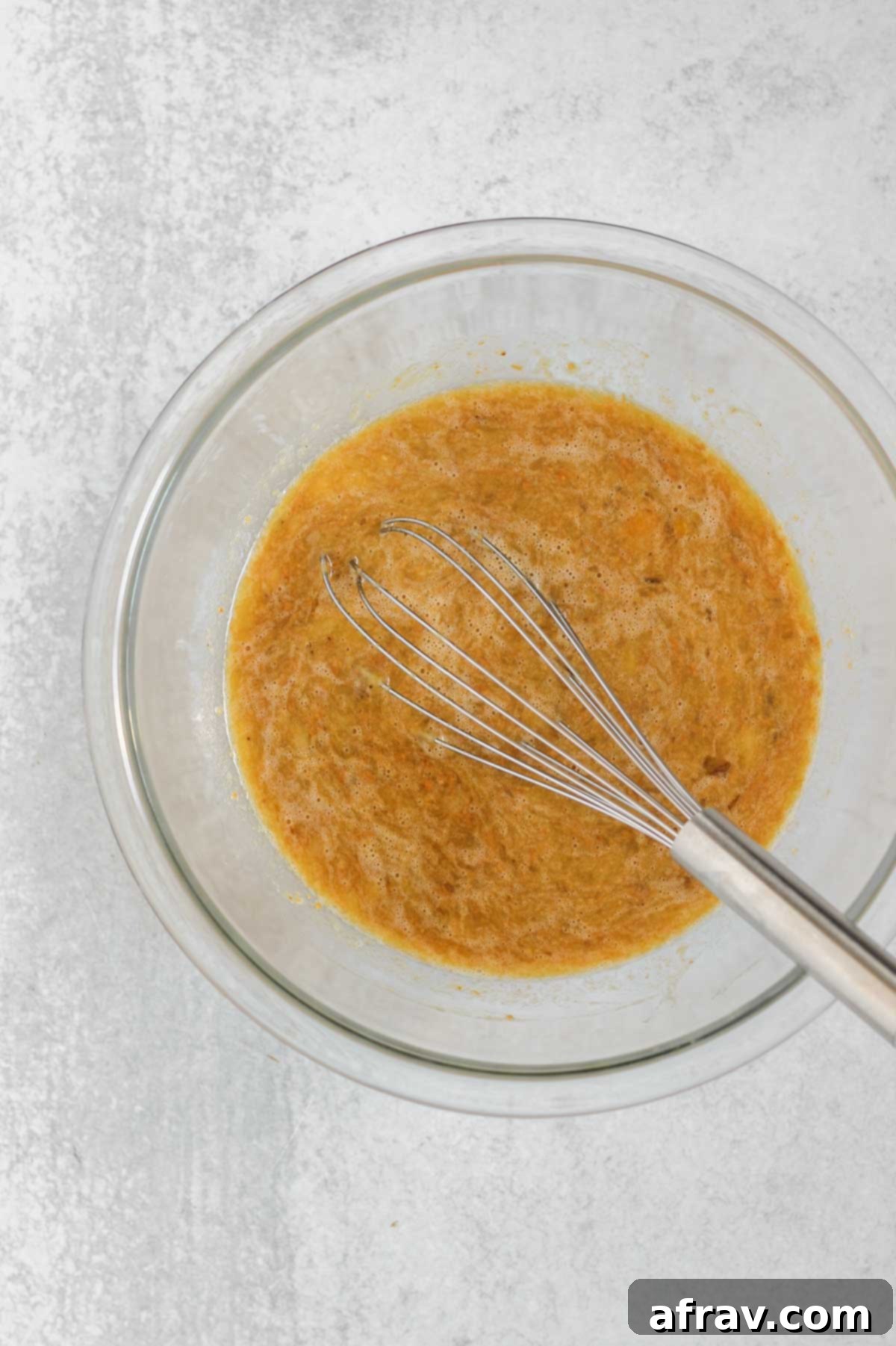 Wet ingredients for muffins, including mashed bananas, eggs, and maple syrup, being whisked together in a clear glass mixing bowl.