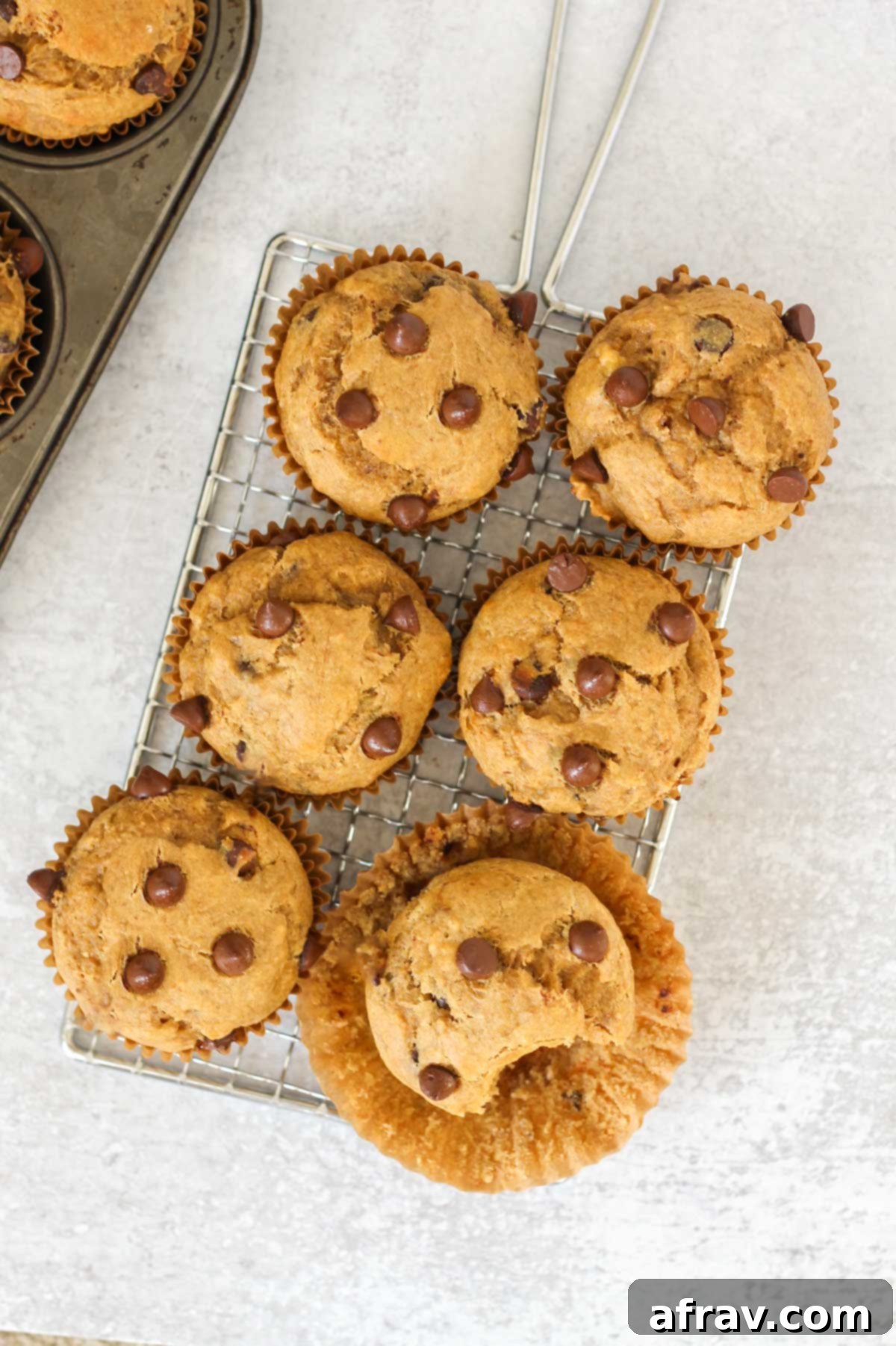 A perfectly baked banana protein muffin with the paper liner peeled back, revealing a soft interior and chocolate chips, with other muffins in the background.