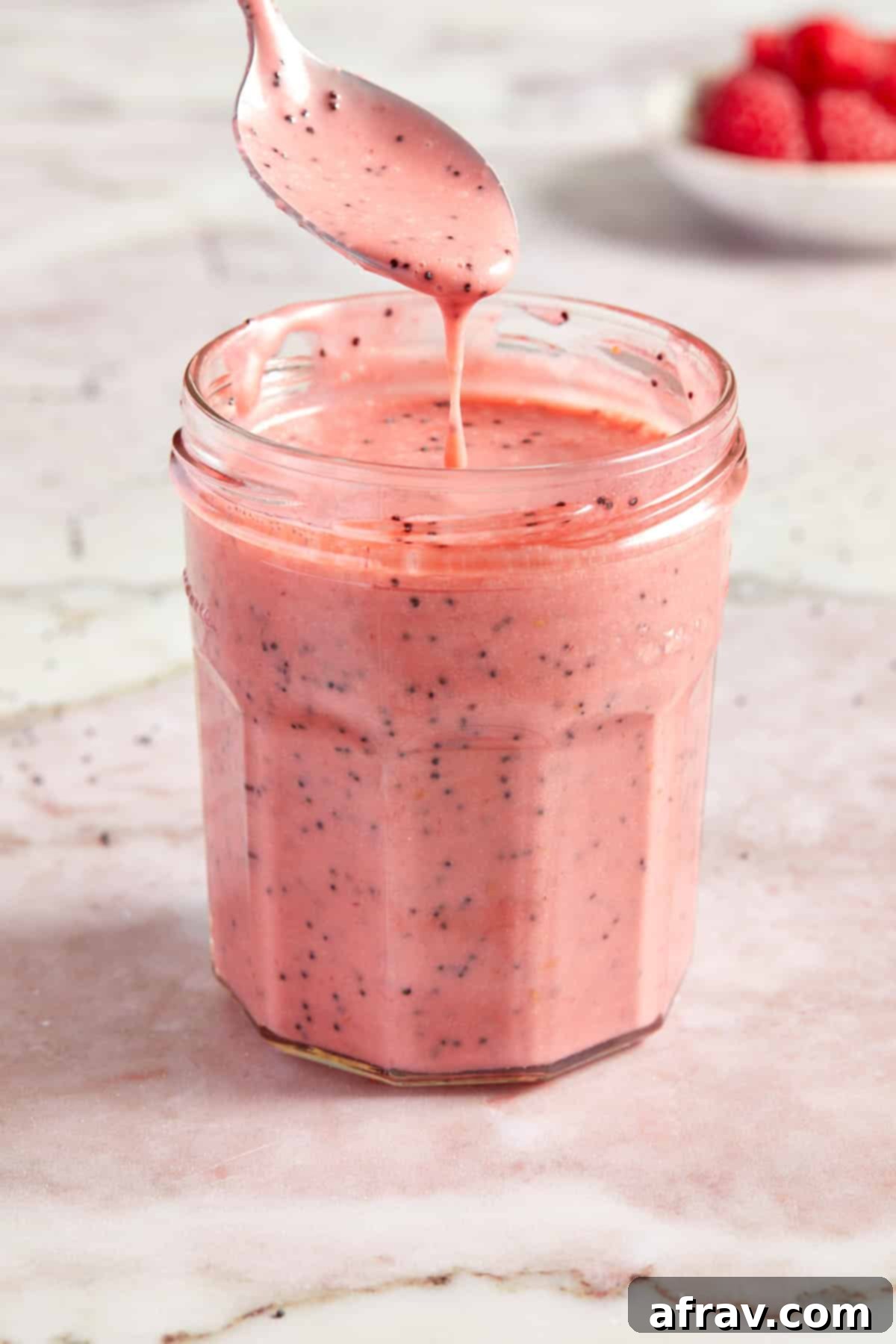 Side view of a glass jar filled with luscious raspberry poppy seed dressing, a spoon delicately lifting a portion, highlighting its thick and smooth consistency.