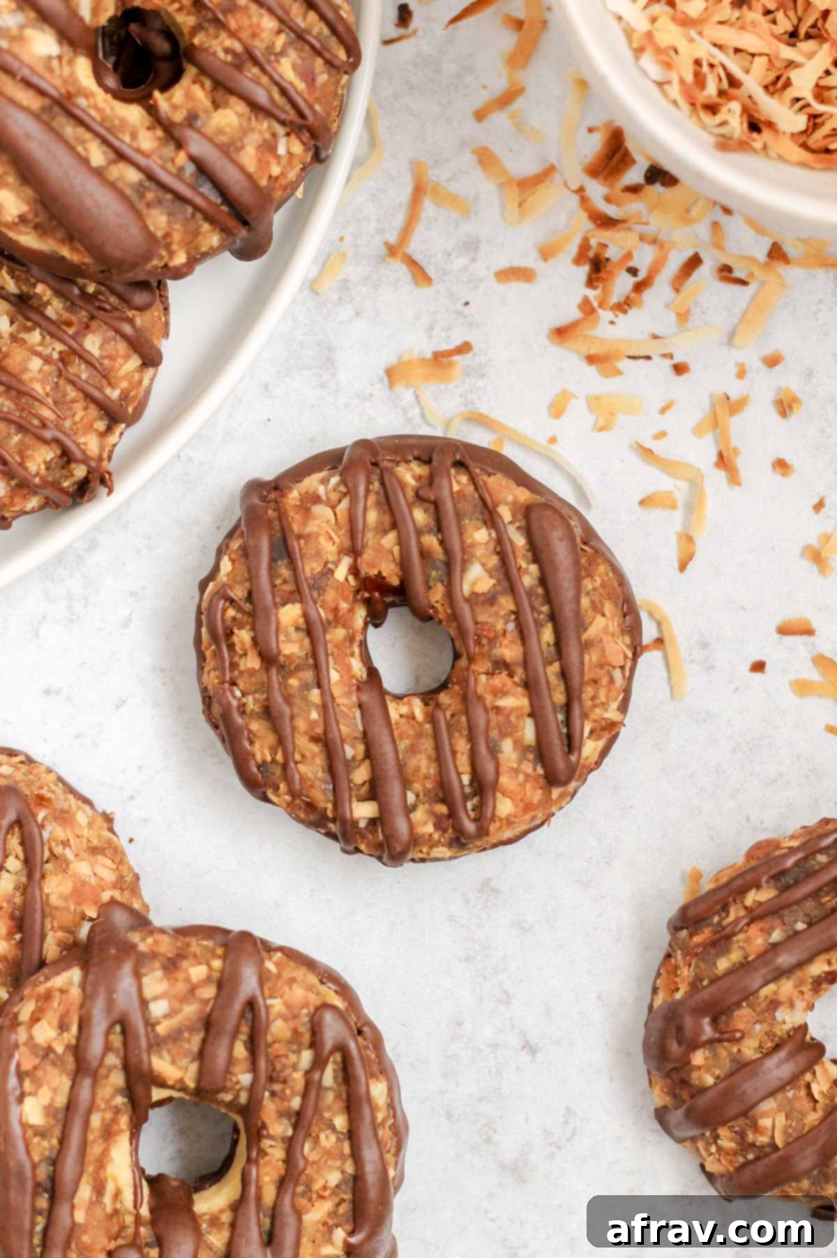 A single healthy Samoa cookie, perfectly crafted with a shortbread base, date caramel coconut topping, and chocolate drizzle, rests on a grey surface. Additional cookies are artfully arranged in the background.