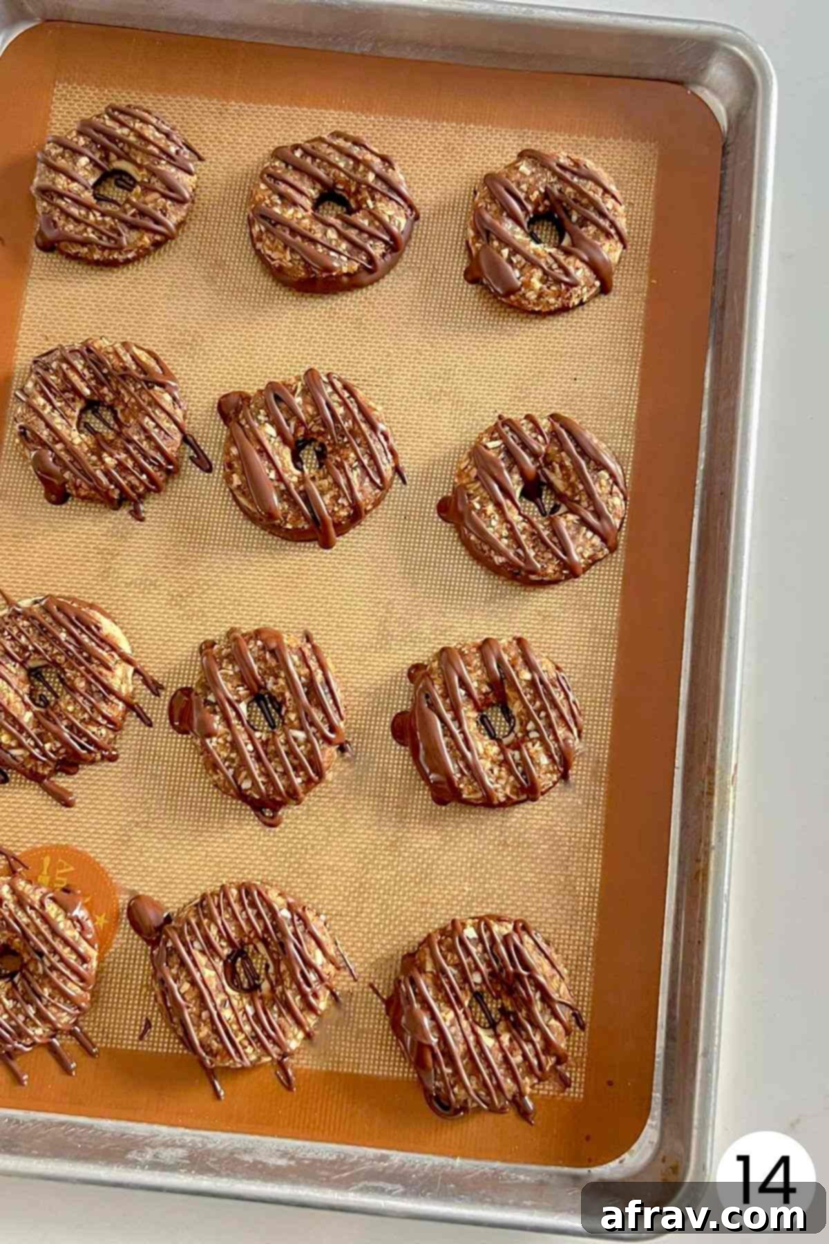 Freshly made Samoa cookies on a baking sheet, with delicate chocolate drizzles on top, numbered 14.