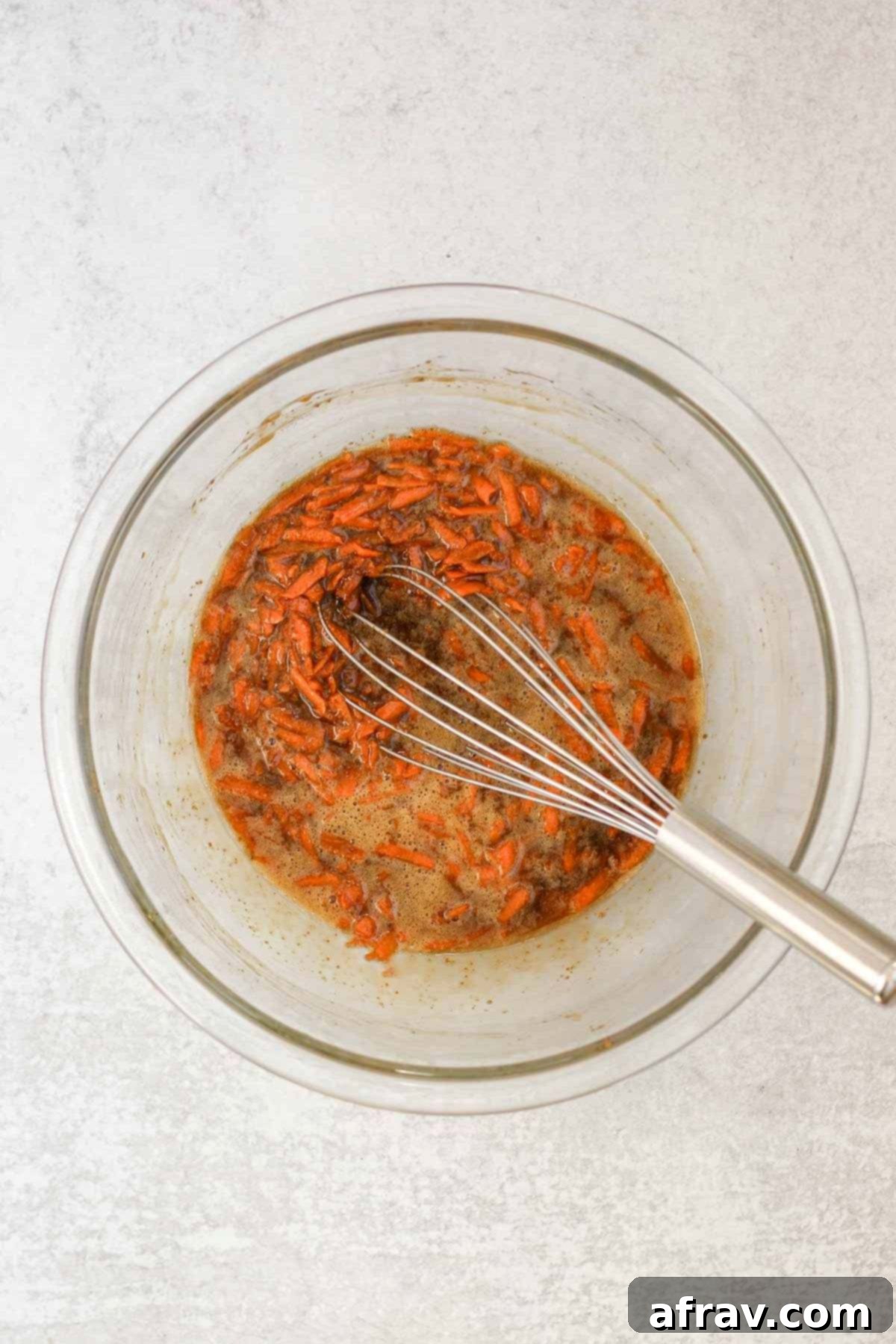 Spiced Carrot Oatmeal Delights 5 Wet ingredients, plus shredded carrots, needed for oatmeal carrot cake cookies in a glass mixing bowl with a wire whisk.