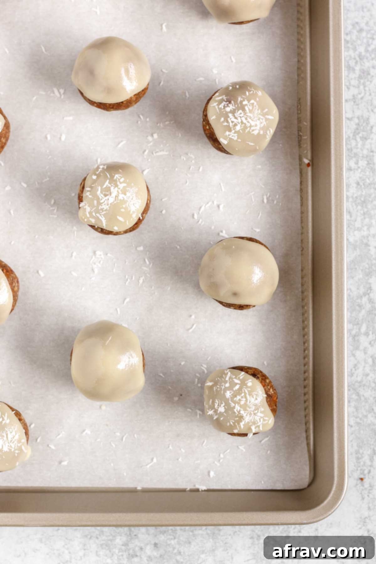 Homemade carrot cake energy balls generously topped with white coconut butter glaze and a sprinkle of coconut flakes, arranged on a baking sheet.