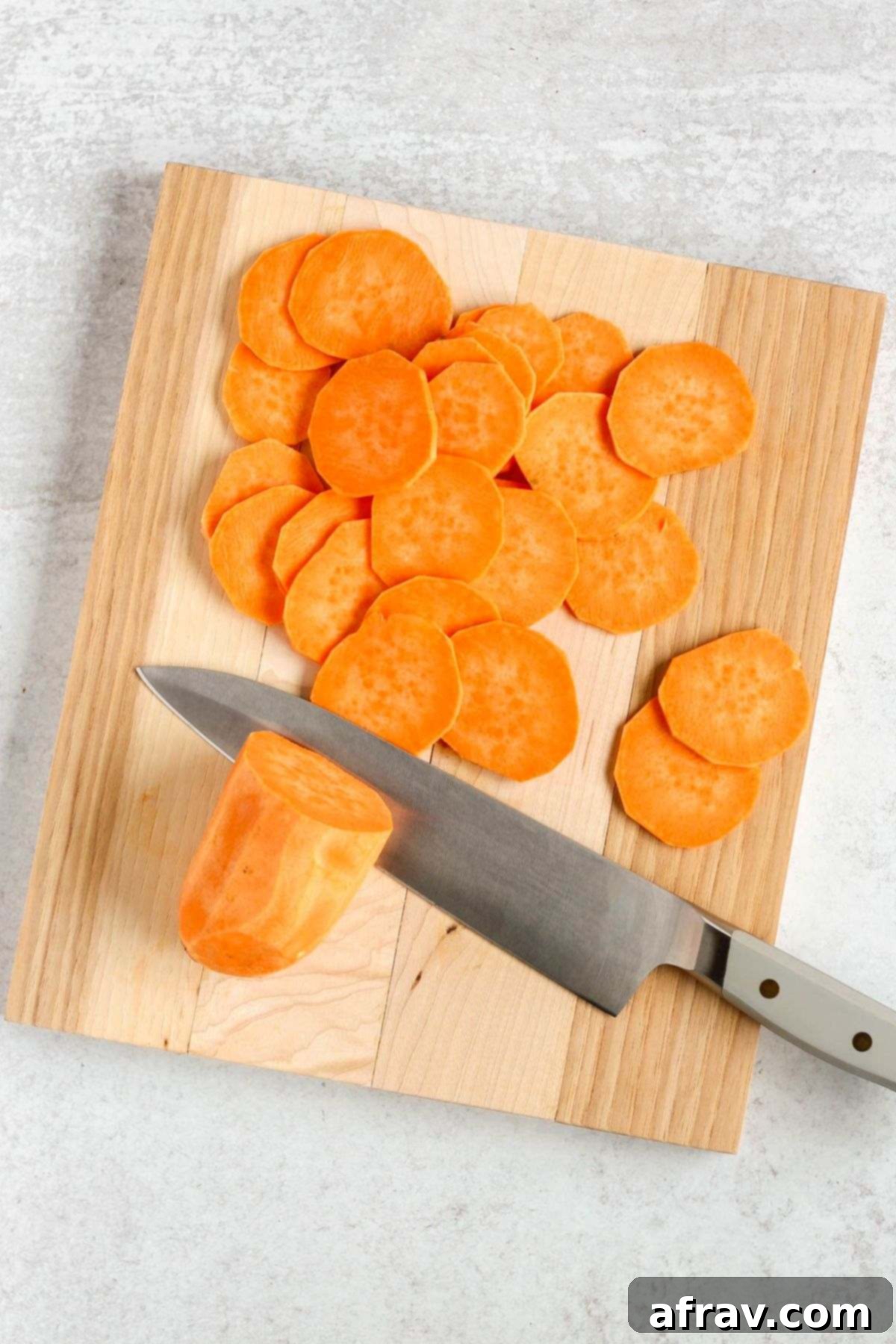 Chef's knife thinly slicing a sweet potato on a wooden cutting board.