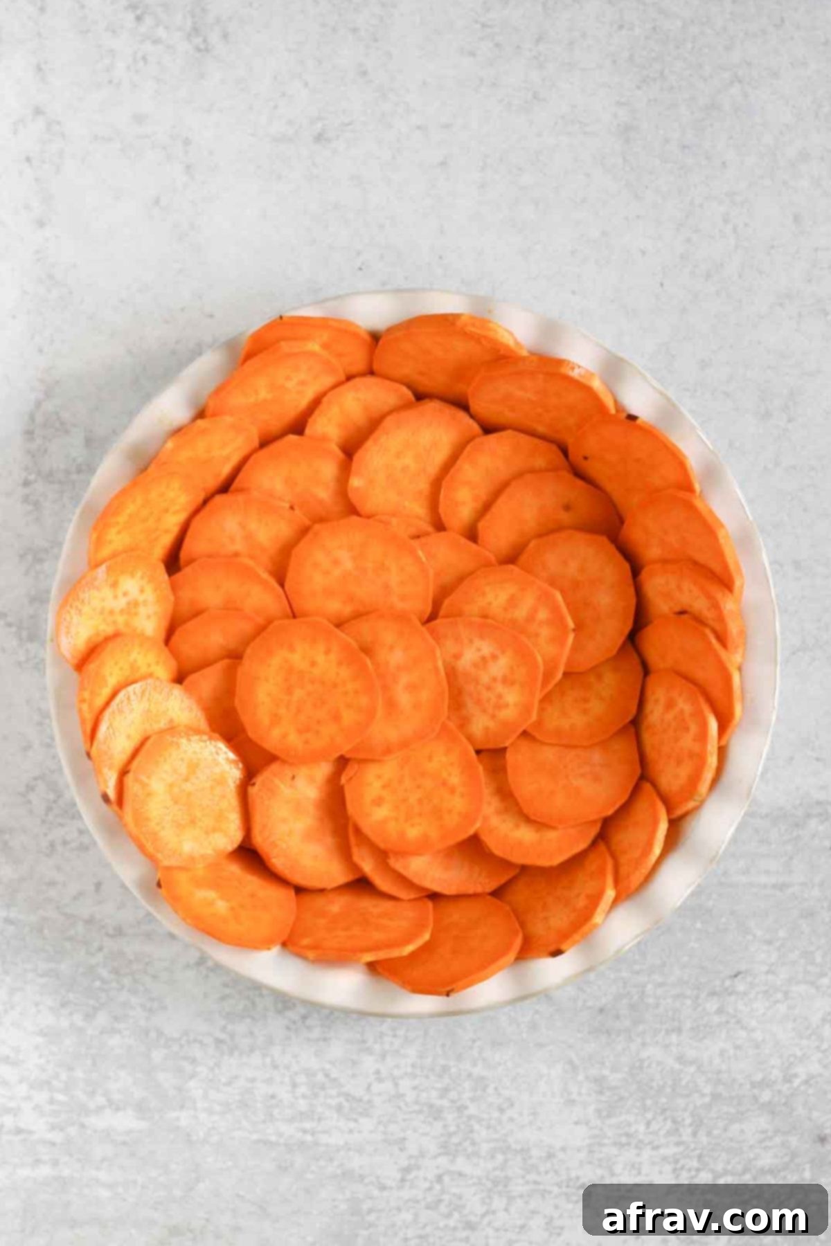 Raw, thinly sliced sweet potatoes arranged in an overlapping pattern in a white ceramic pie dish, ready for baking.