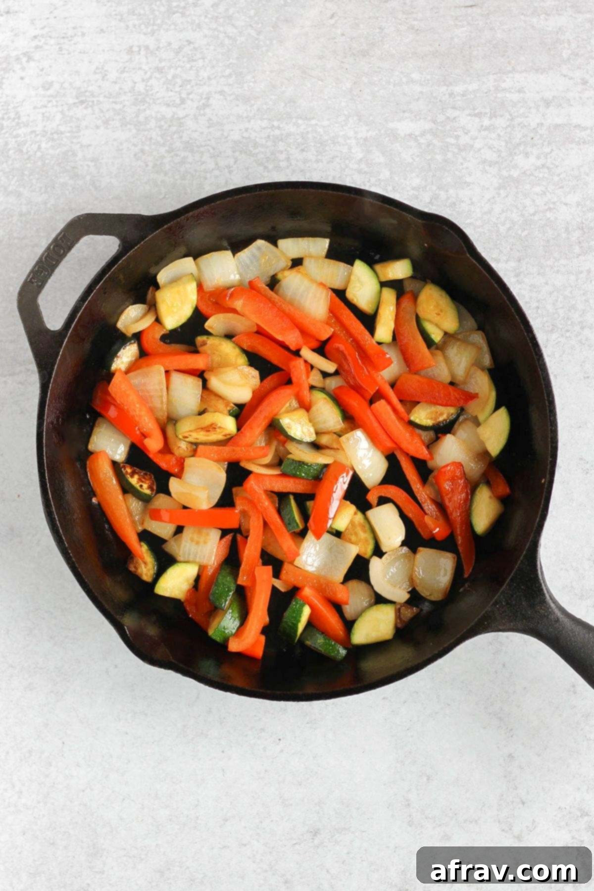 Sautéed red peppers, zucchini, and yellow onion cooking in a cast iron skillet, softening and becoming fragrant.