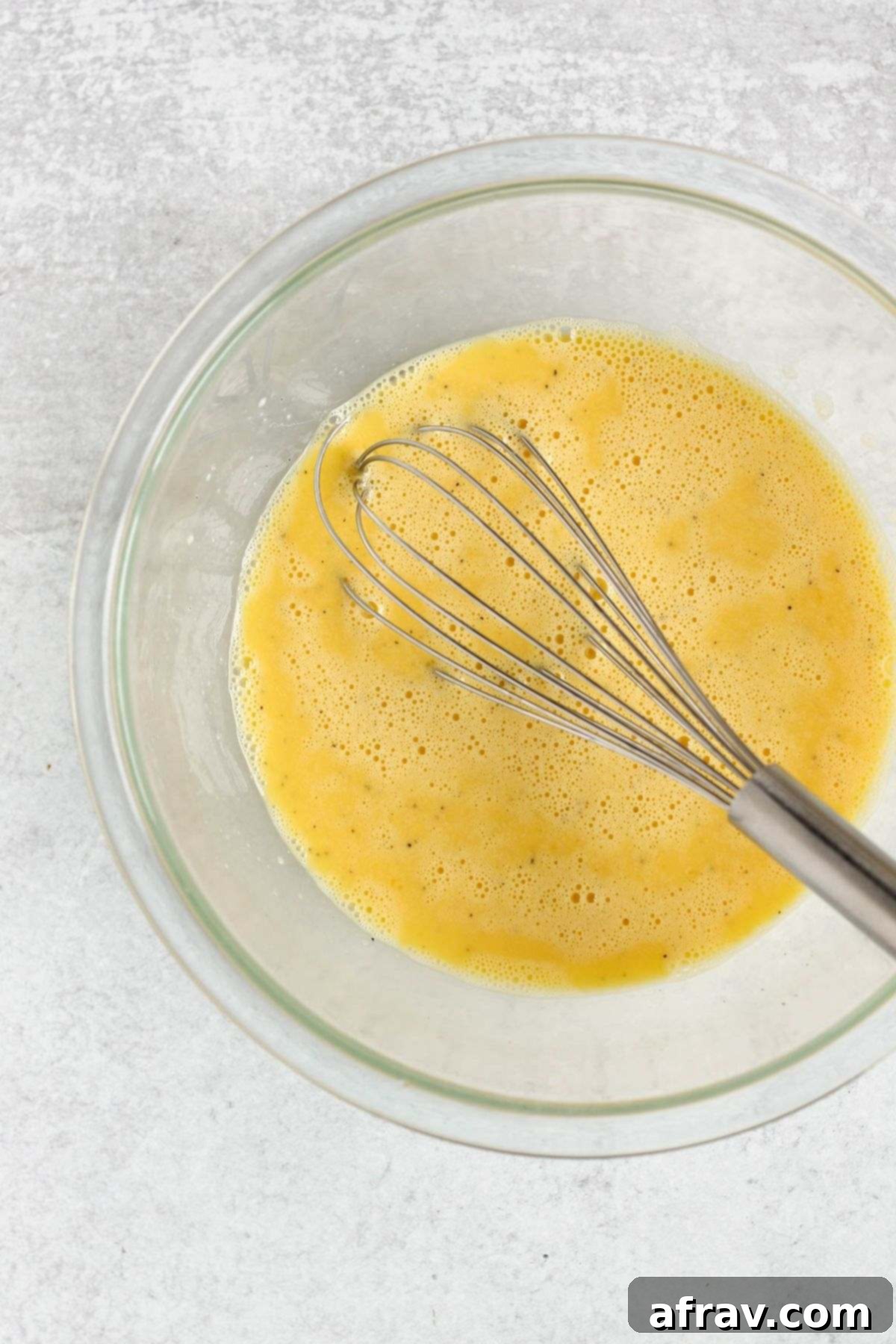 Whisked eggs in a clear glass mixing bowl, light and frothy, with a wire whisk resting in the bowl.
