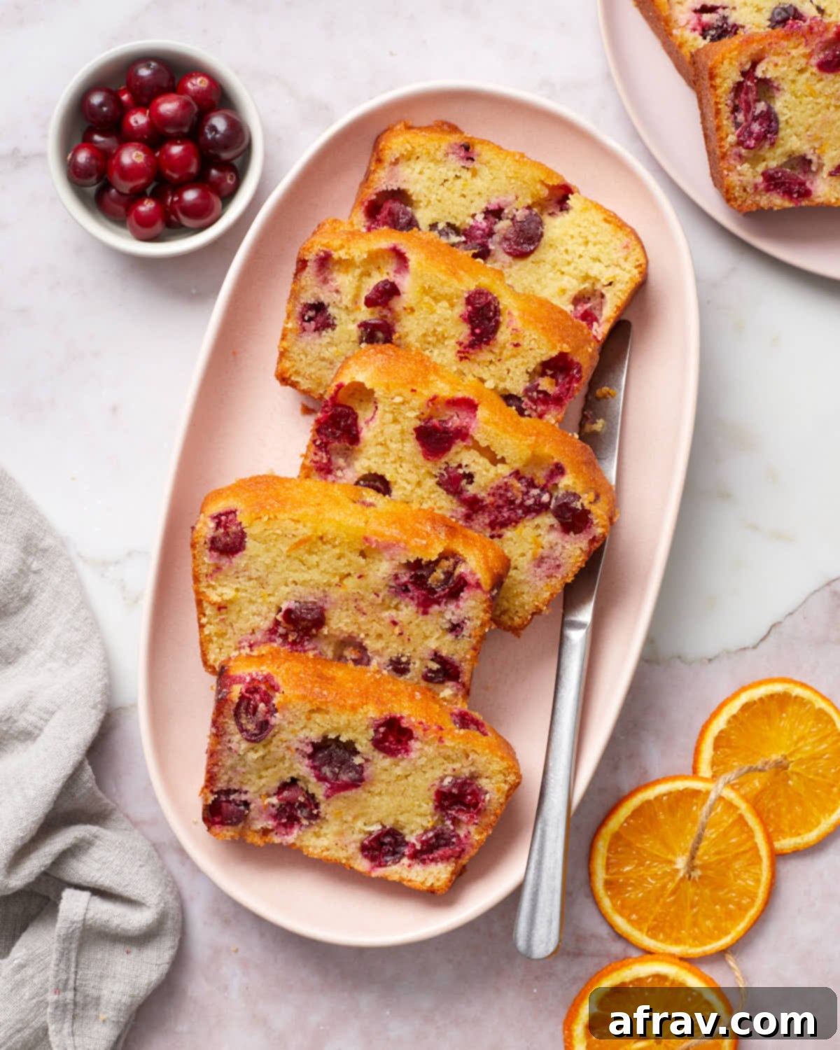 Slices of cranberry orange bread arranged on a pink platter with cranberries and orange slices to the side, highlighting the moist texture and fruit.