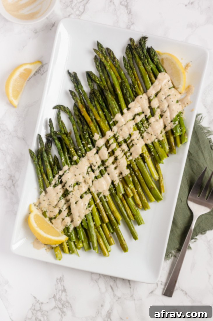 Easy Roasted Asparagus with Creamy Lemon Tahini Sauce 2 Perfectly roasted asparagus spears drizzled generously with a creamy lemon tahini sauce on a white serving plate, ready to be enjoyed.