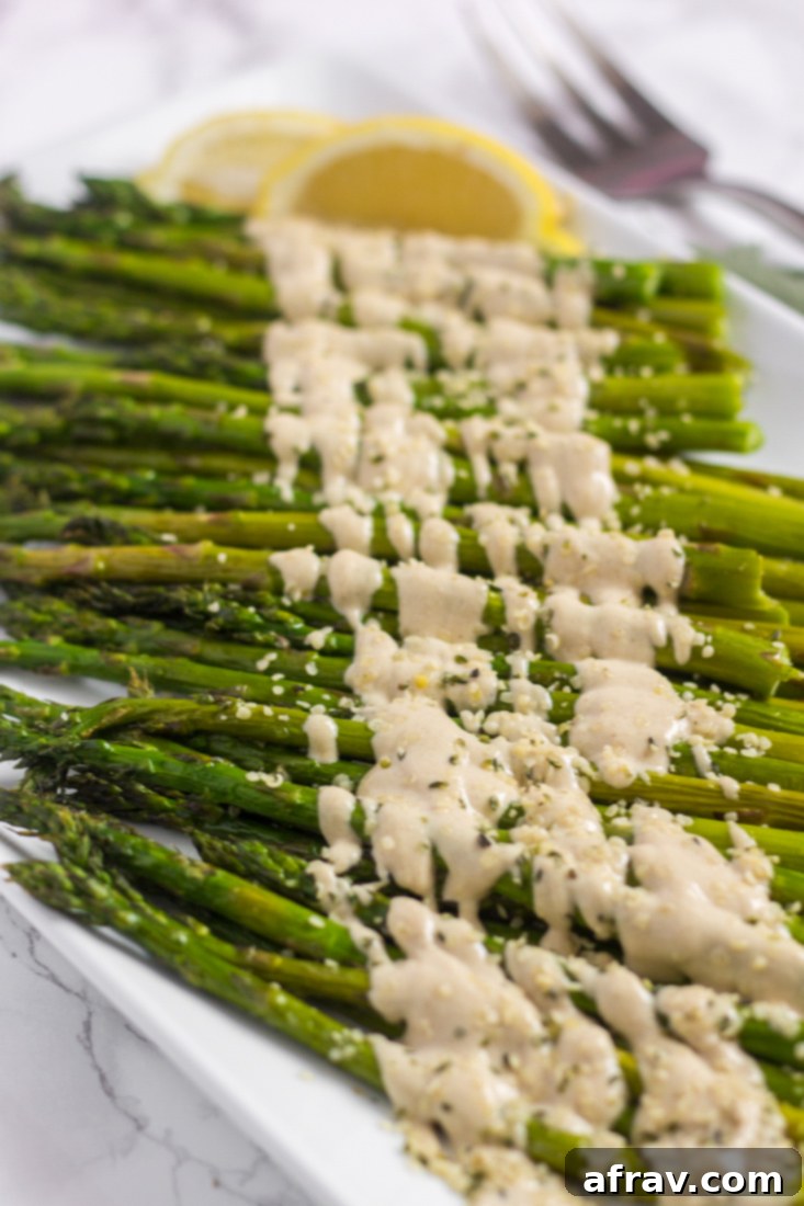 Easy Roasted Asparagus with Creamy Lemon Tahini Sauce 5 Close-up side view of roasted asparagus spears generously drizzled with a creamy lemon tahini sauce on a white plate.