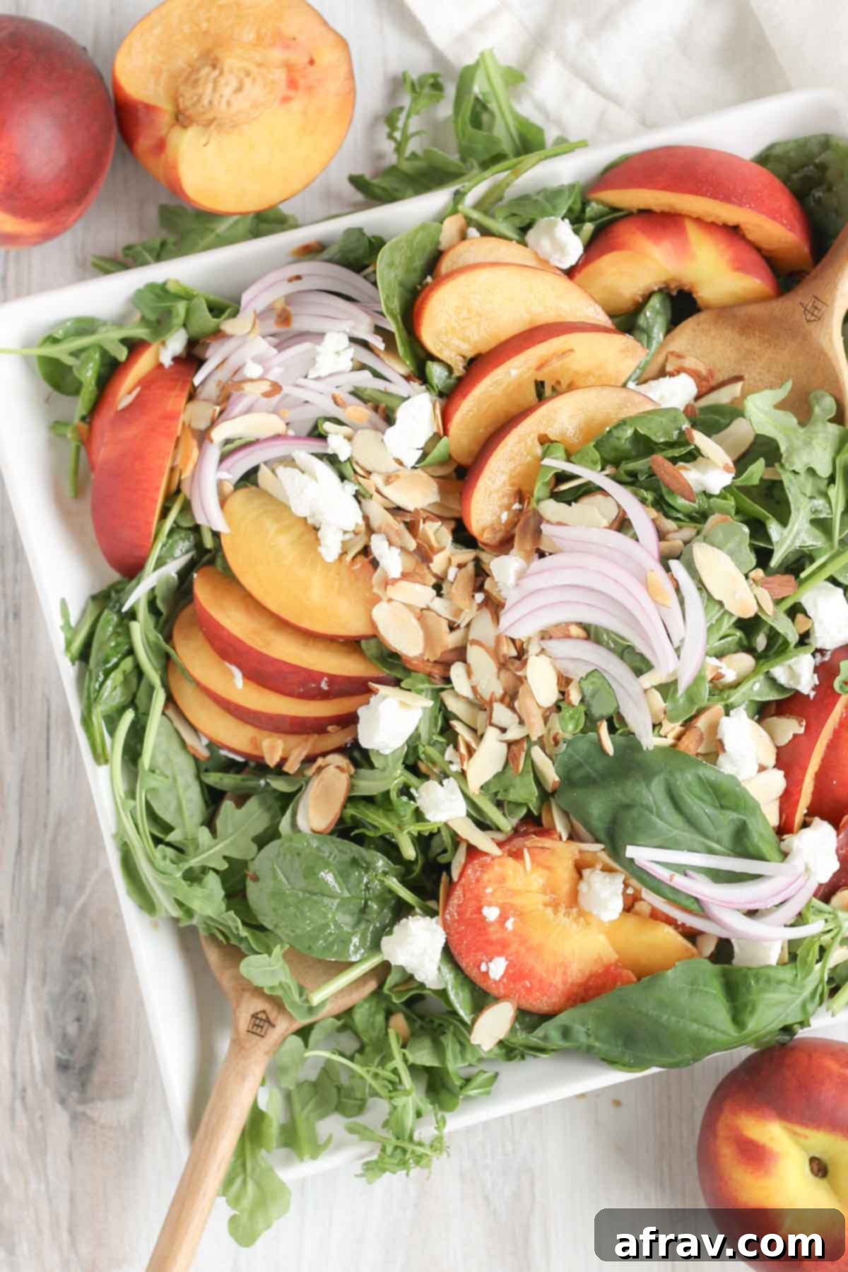 Wooden salad tongs mixing together a peach and goat cheese salad on a square plate with peaches on the sides, ready to be served.