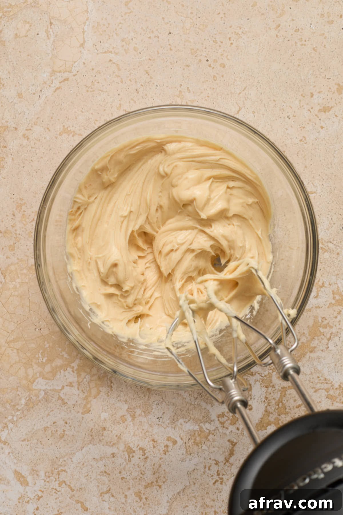 Frosting ingredients being whipped together in a glass bowl with electric beaters.