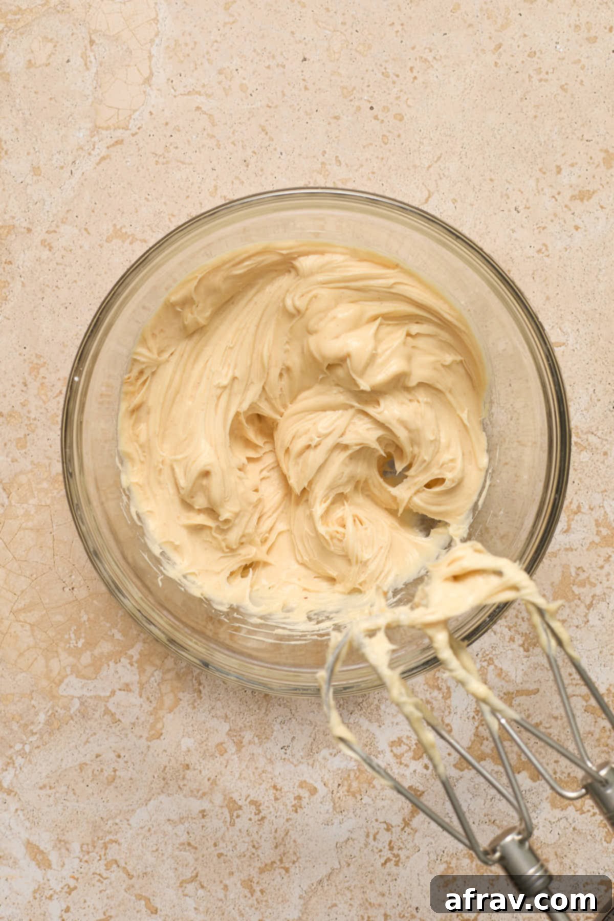 Close-up of freshly made peanut butter cream cheese frosting, light brown and smooth, in a clear glass mixing bowl.