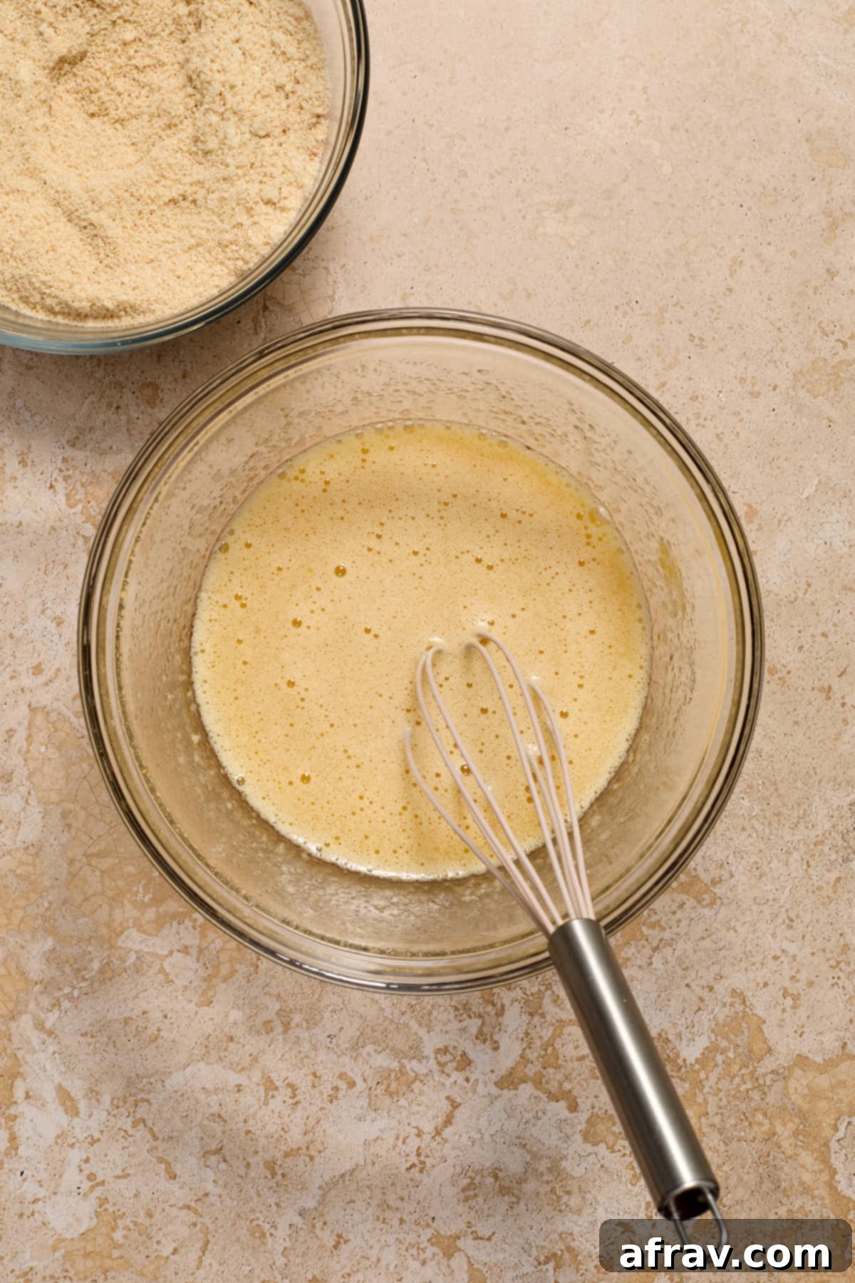Wet ingredients for the cake, including eggs, sugar, oil, applesauce, and vanilla, whisked in a bowl with dry ingredients nearby.