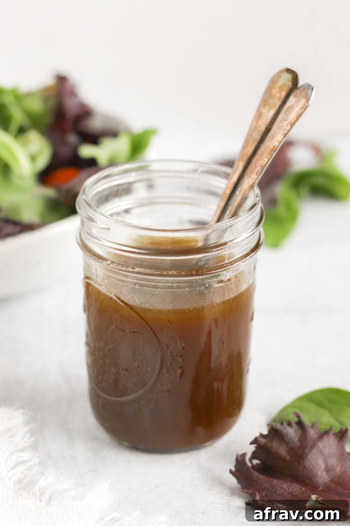 Effortless Balsamic Vinaigrette 5 Mason jar with two spoons filled with balsamic vinaigrette with a bowl of mixed greens in the background.