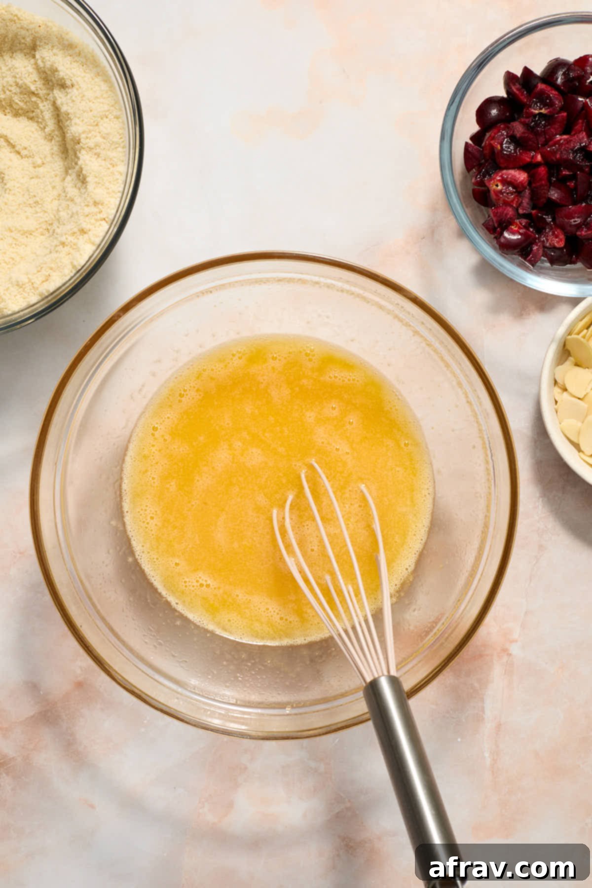 Wet ingredients whisked together in a bowl with dry ingredients, almonds and cherry surrounding it.
