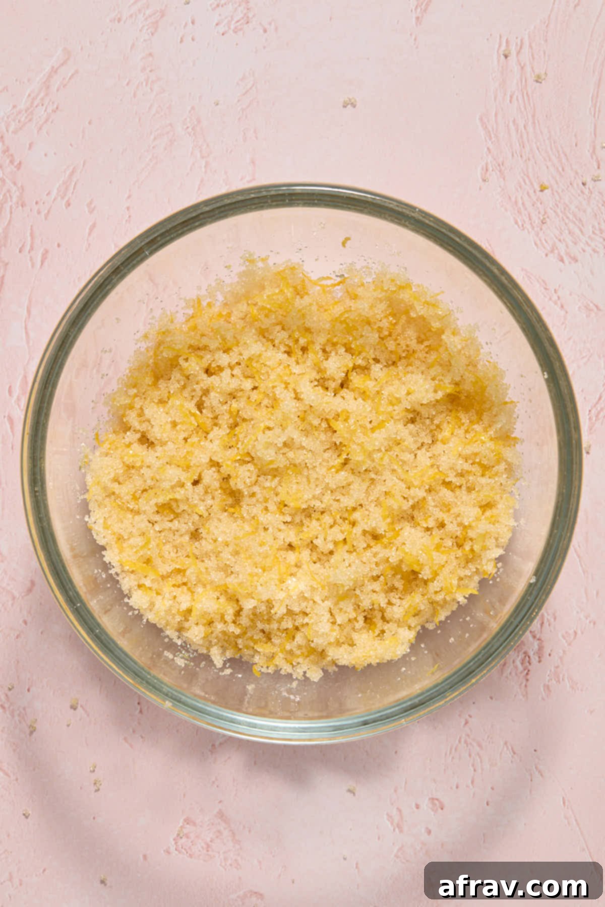 Lemon zest and cane sugar being rubbed together between fingers in a small glass bowl, appearing wet and fragrant.