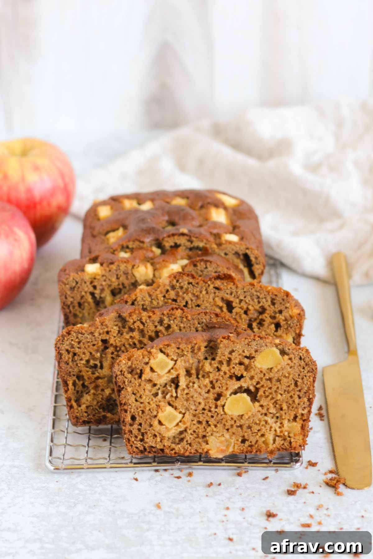 Multiple slices of healthy apple bread neatly arranged on a rectangular wire cooling rack with a golden knife on the side and fresh apples in the background.