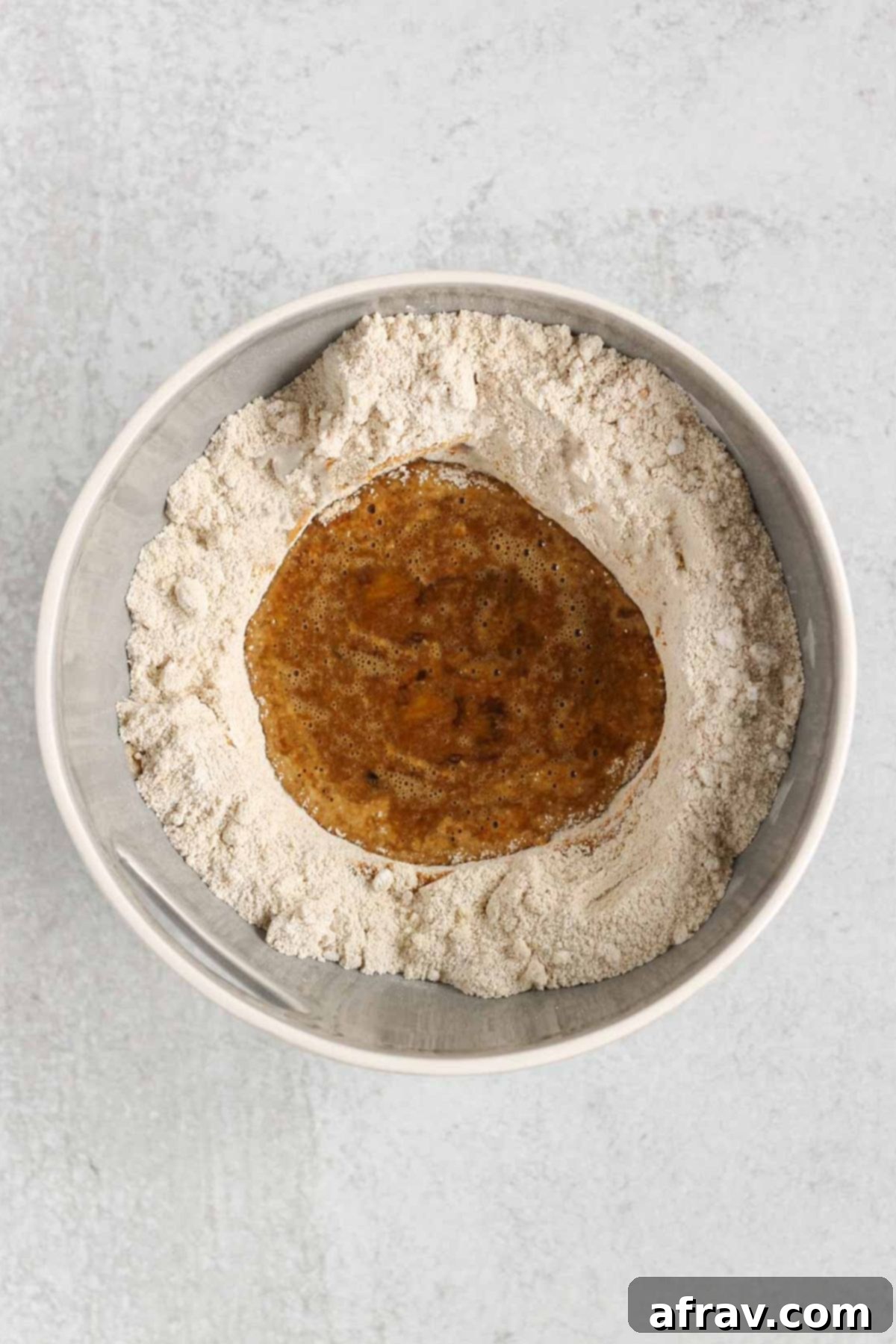 Wet ingredients, including eggs and applesauce, poured into the well of the dry flour mixture in a mixing bowl for apple bread batter.