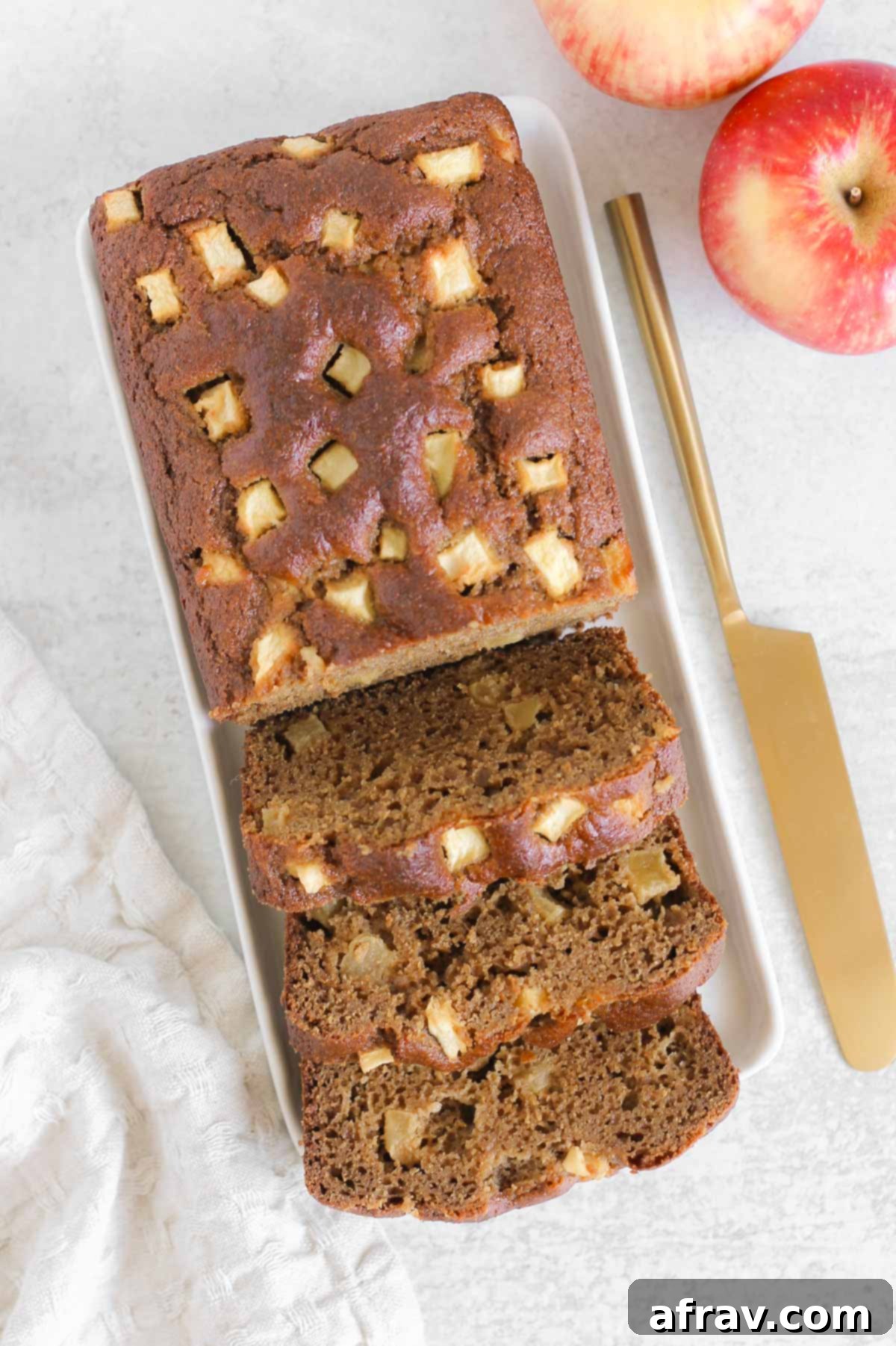 Half sliced loaf of healthy apple bread on a rectangular plate with a gold knife and fresh apples on the side, ready to be enjoyed as a wholesome treat.
