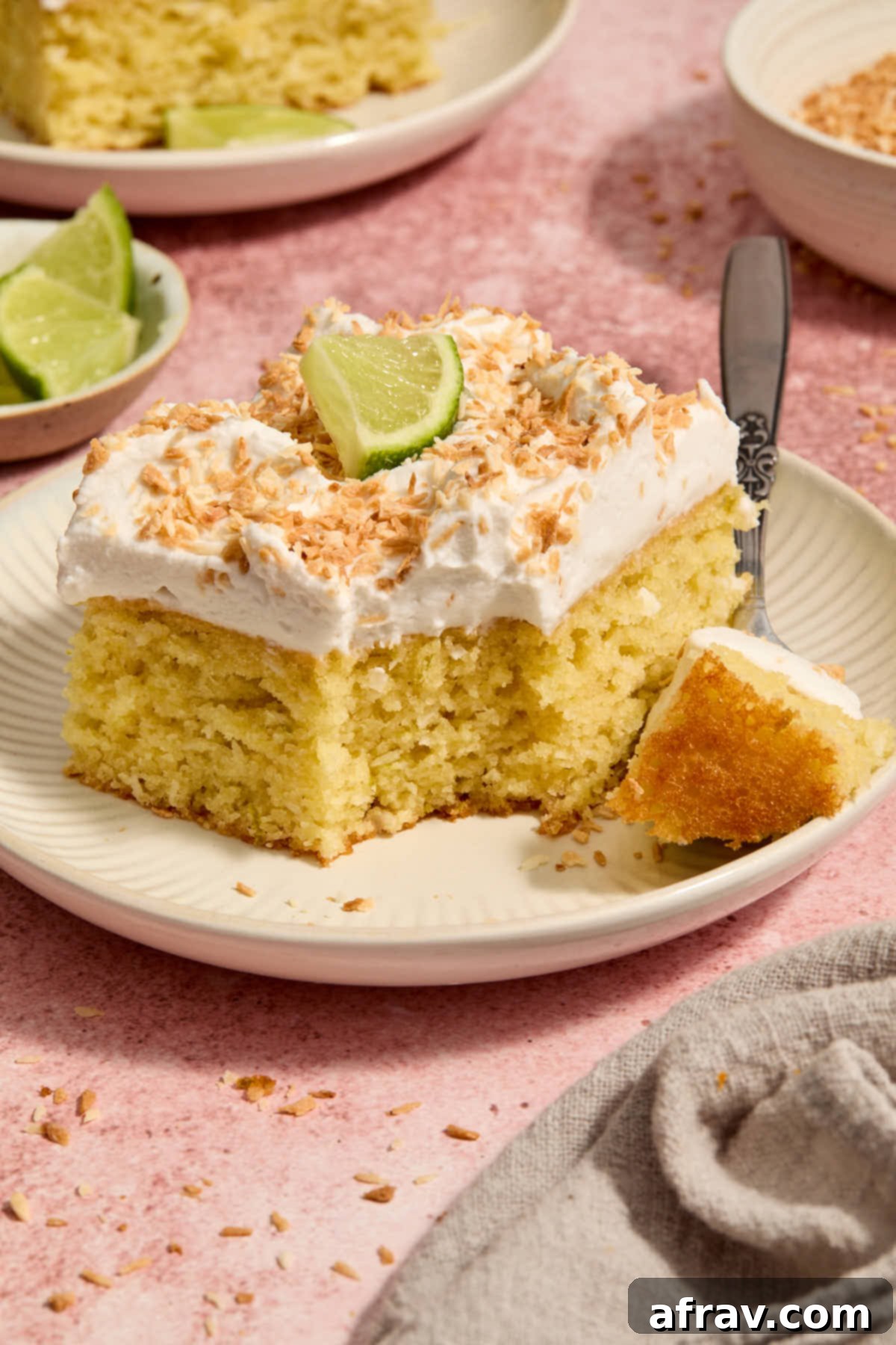 Close-up side view of a slice of almond flour coconut lime cake, garnished with a fresh lime slice and showing a forkful removed, emphasizing its moist interior.