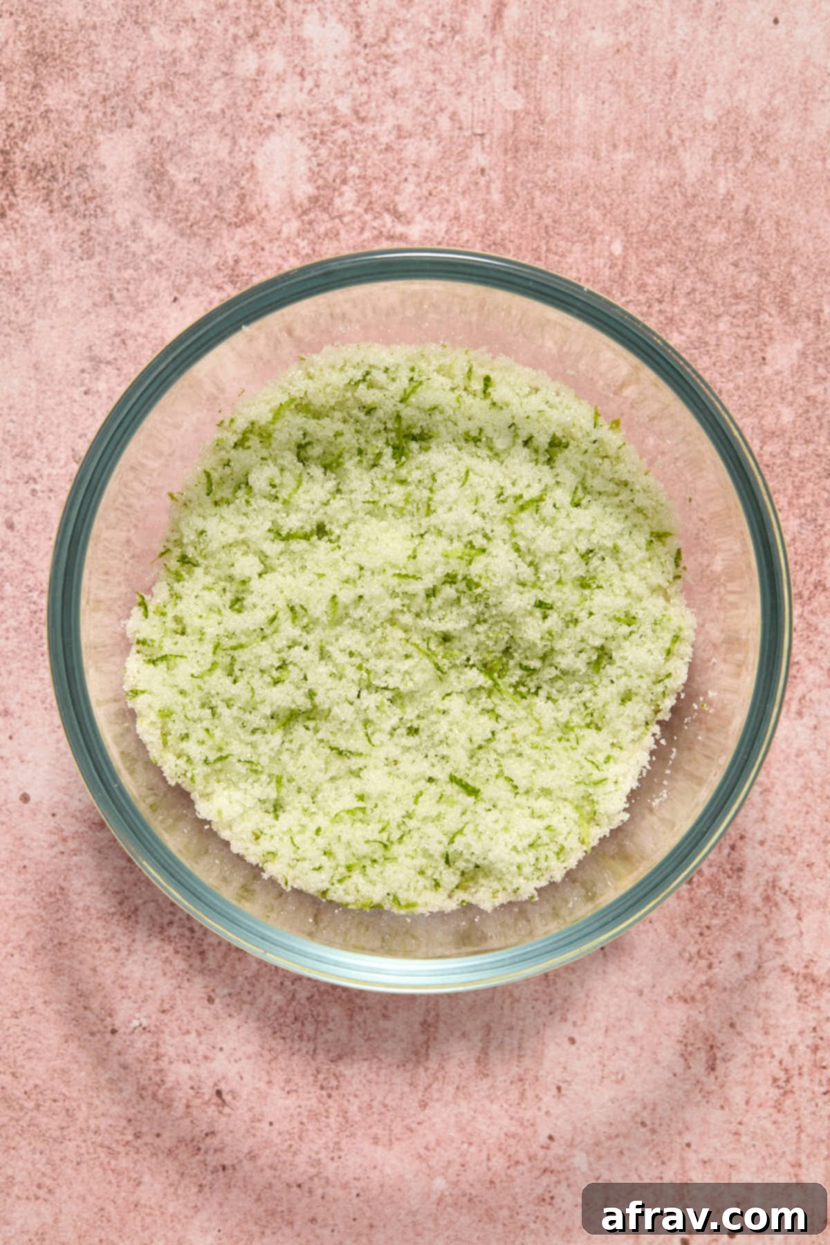 Granulated sugar and fresh lime zest being rubbed together between fingers in a small glass bowl to release aromatic oils.