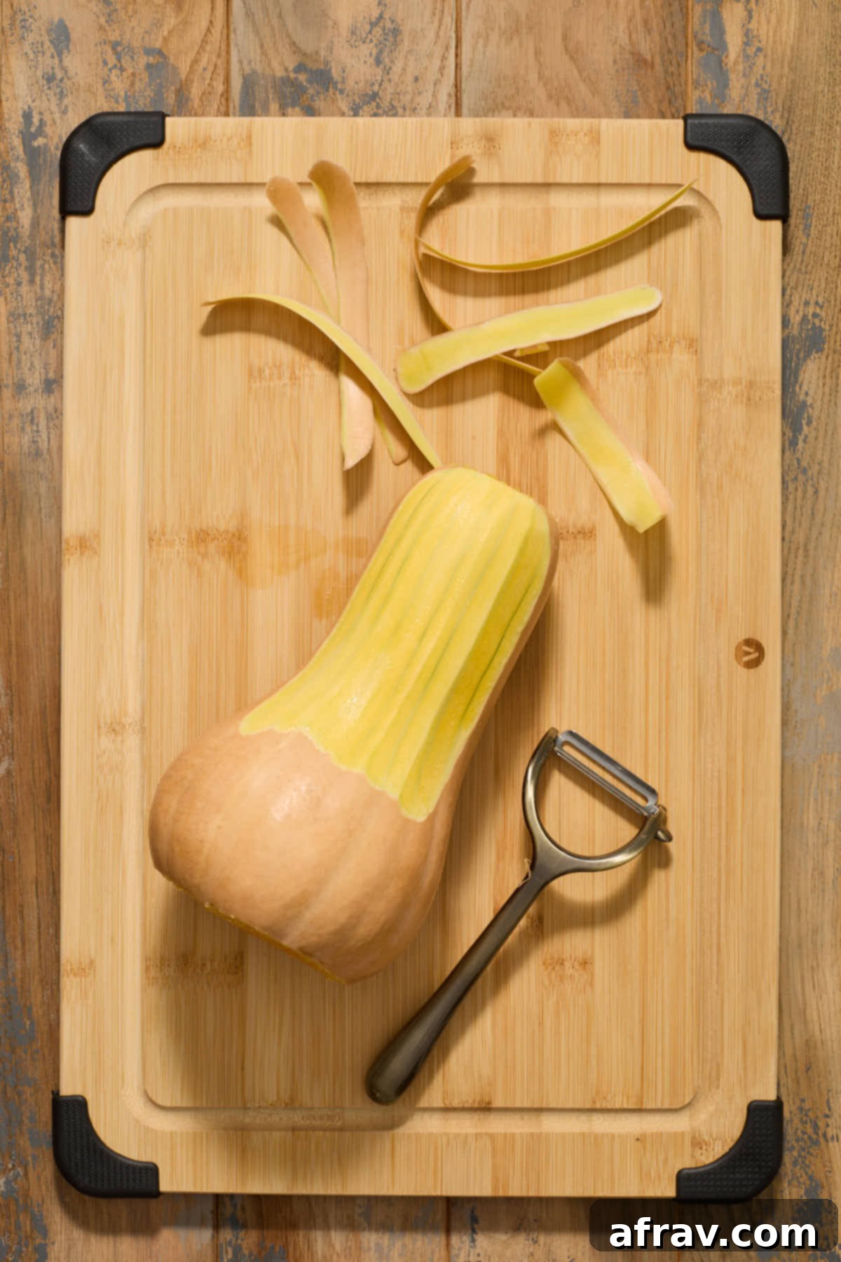 The elongated neck portion of a butternut squash being peeled downwards with a Y-peeler on a wooden cutting board, revealing bright orange flesh.