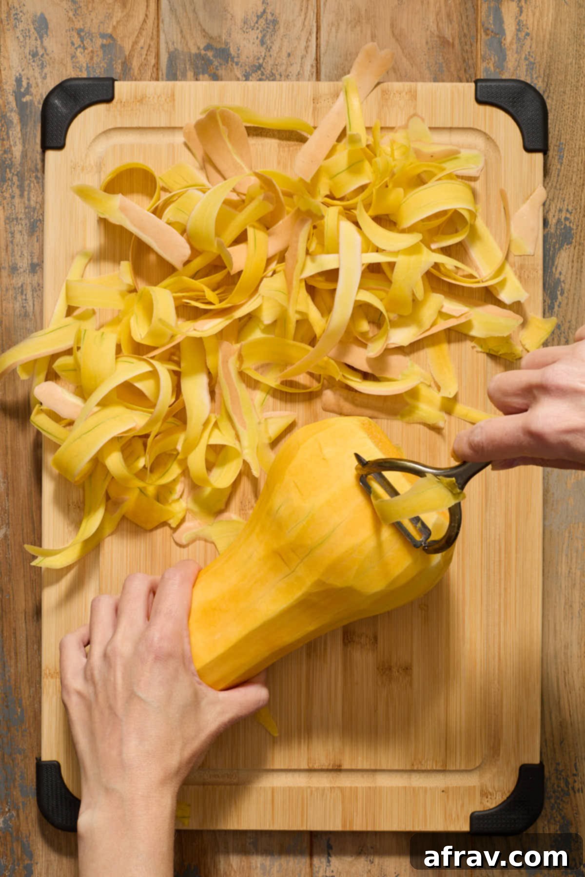 A fully peeled butternut squash resting on a wooden cutting board, surrounded by ribbons of discarded skin, ready for the next cutting step.