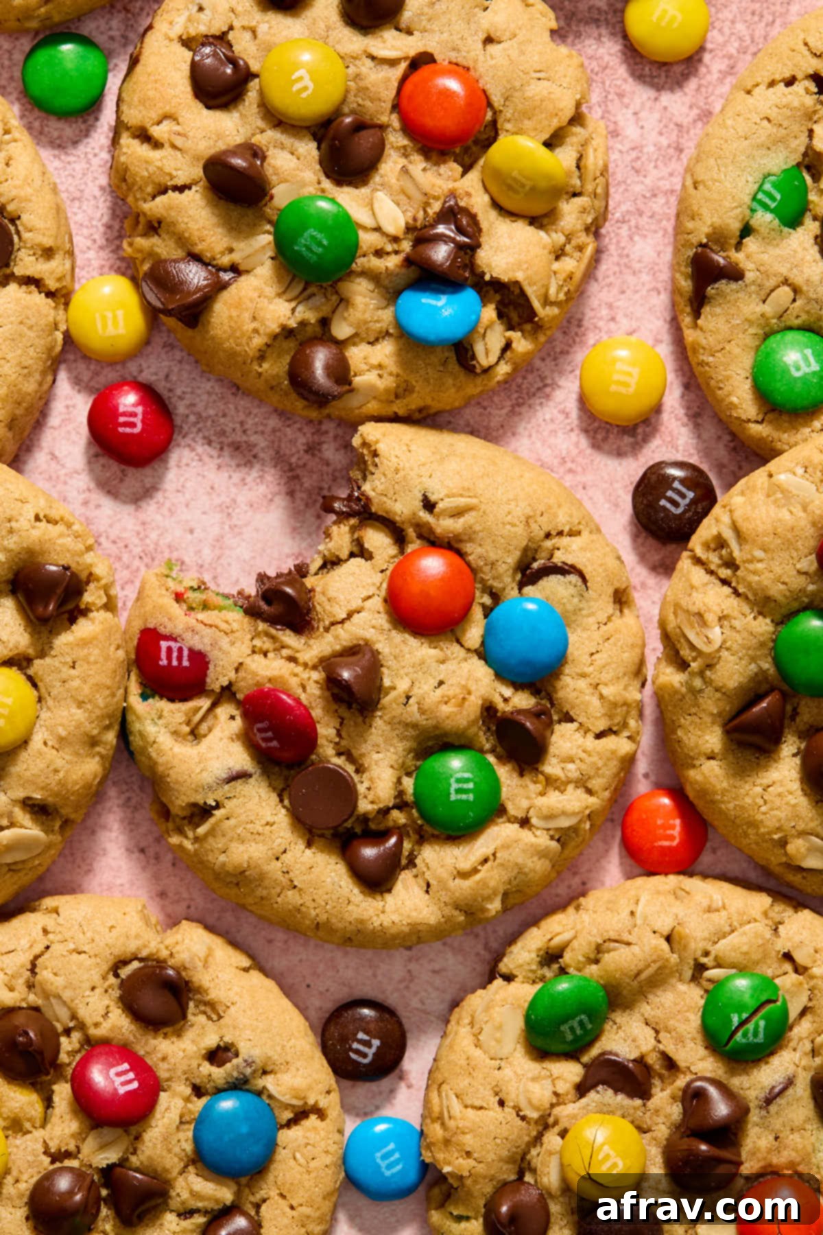 Monster cookies arranged on a pink surface with a bite taken out of one, revealing its soft interior and colorful M&Ms.