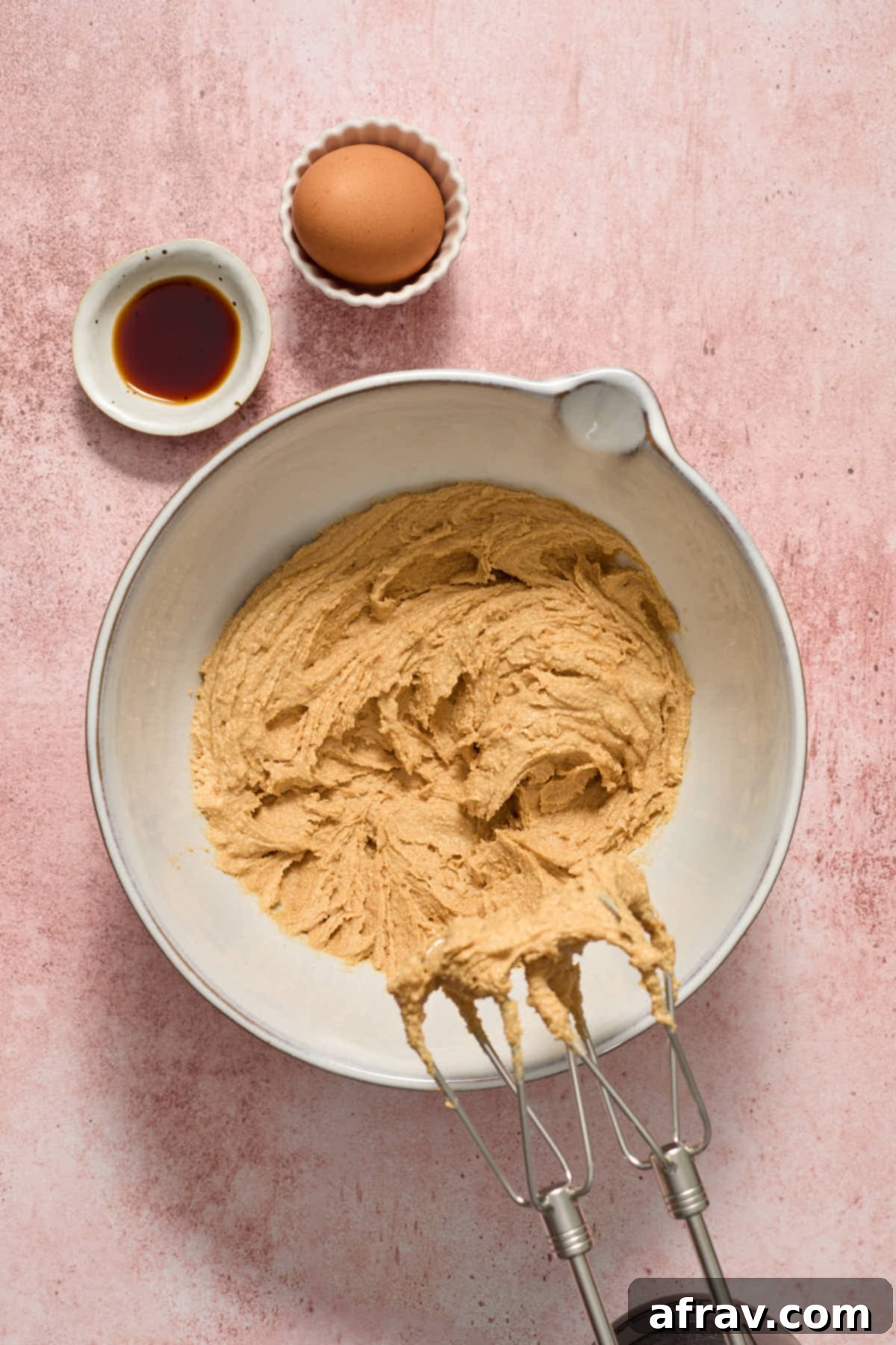 Peanut butter, butter, and sugars creamed together in a bowl with an electric mixer. An egg and vanilla are set aside.