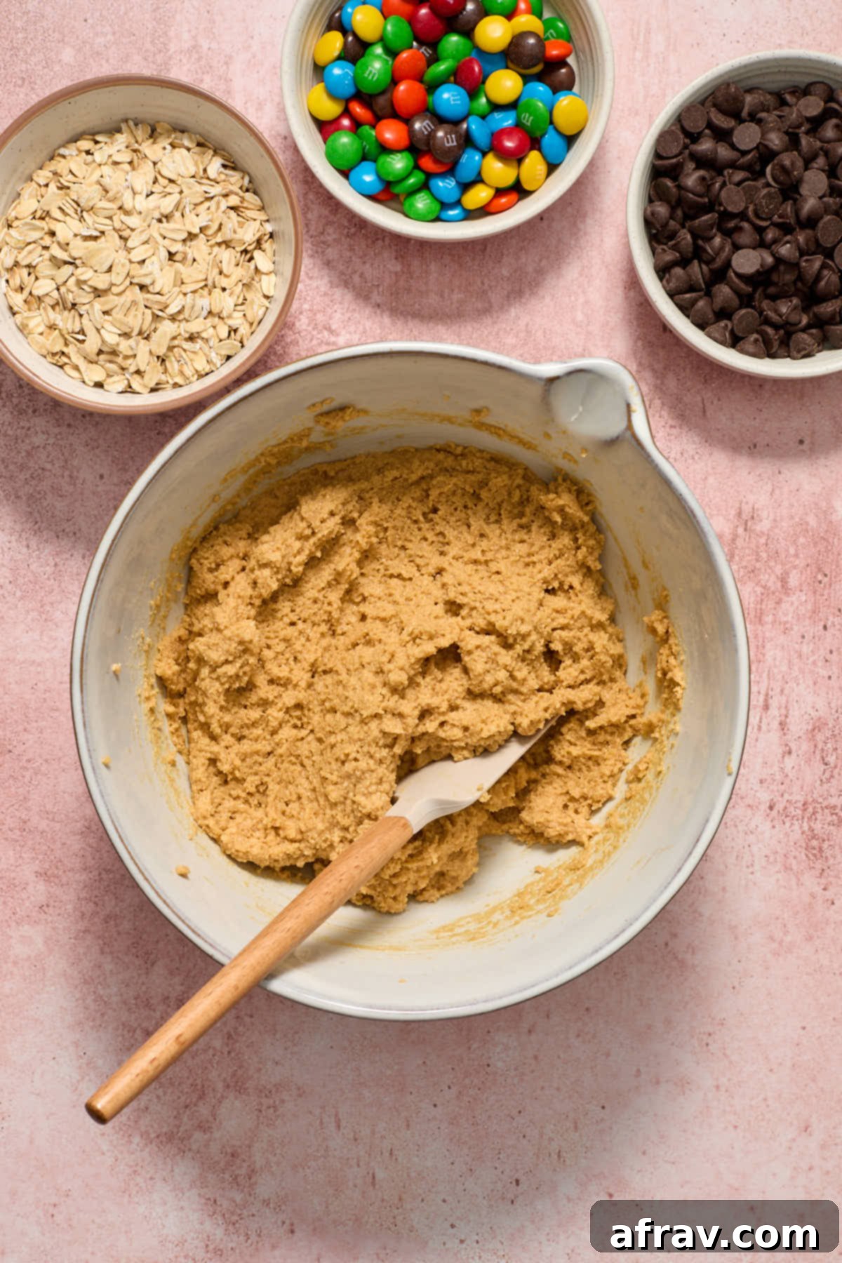 Cookie dough in a bowl after dry ingredients have been added, with separate bowls of oats, M&Ms, and chocolate chips on the side, ready to be mixed in.