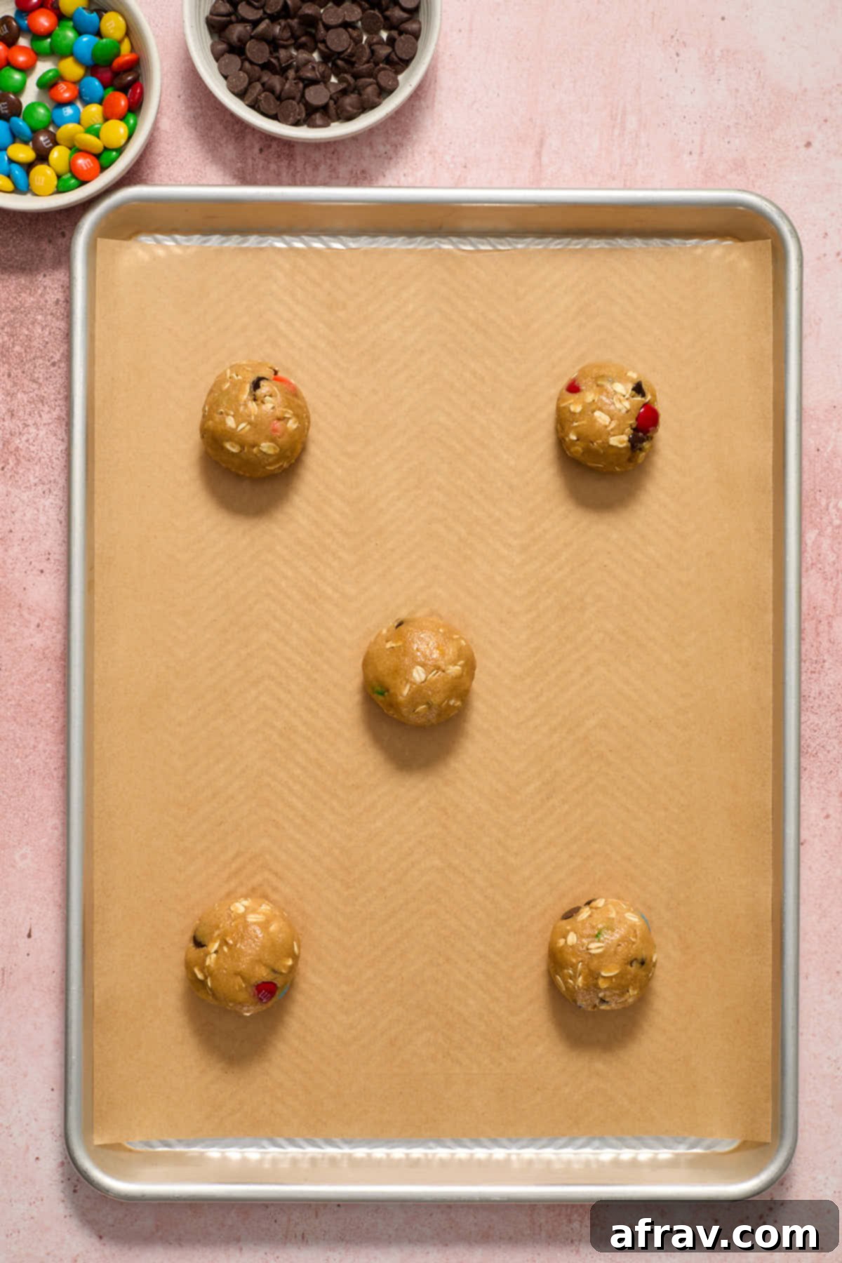 Cookie dough balls arranged on a parchment-lined baking sheet, with additional M&Ms and chocolate chips on the side for topping.