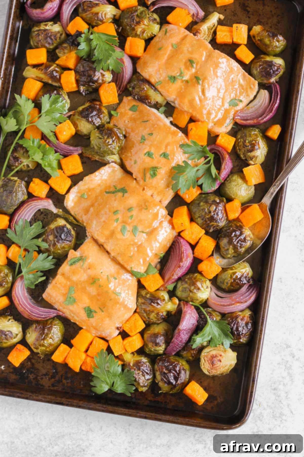 Sheet Pan Maple Dijon Salmon Dinner 8 Roasted salmon and veggies on a baking sheet with parsley sprinkled around and a vintage spoon scooping up some of the veggies.