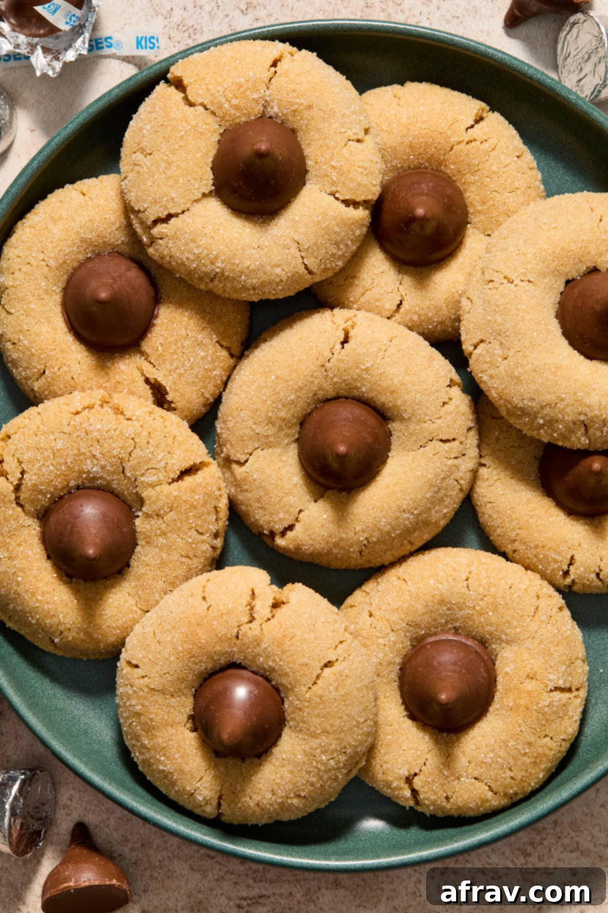 Overhead shot of freshly baked Almond Flour Peanut Butter Blossoms piled on a vibrant green plate, with shiny unwrapped Hershey Kisses scattered around.
