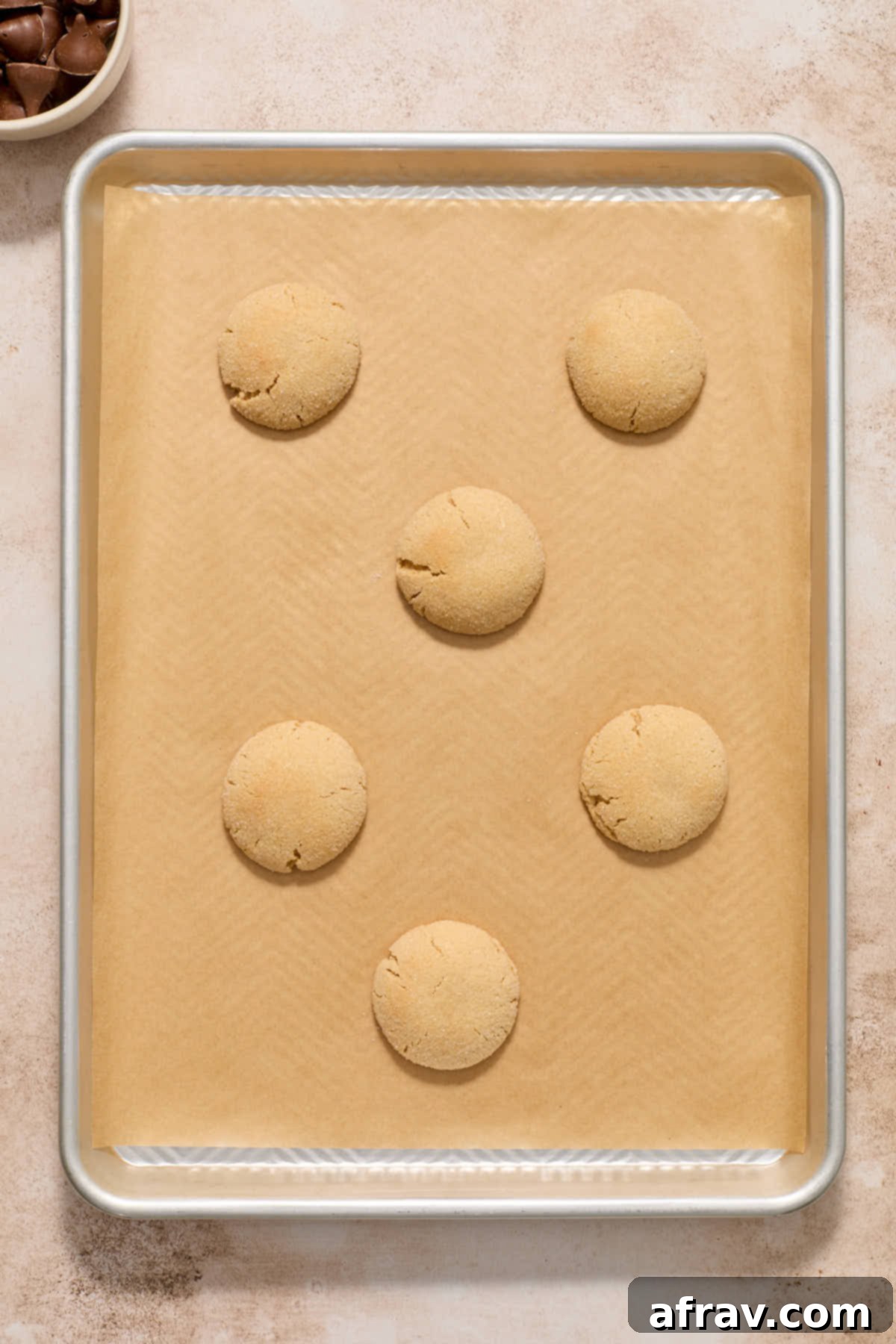 A baking sheet filled with golden-brown, puffed-up peanut butter blossom cookies fresh out of the oven, with a bowl of unwrapped chocolate kisses ready to be pressed.