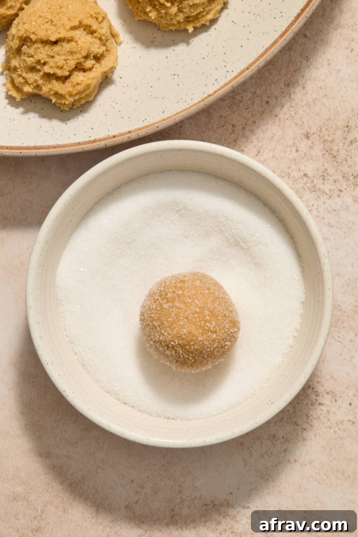A chilled peanut butter cookie dough ball being gently rolled in granulated sugar, ensuring a full and even coating.