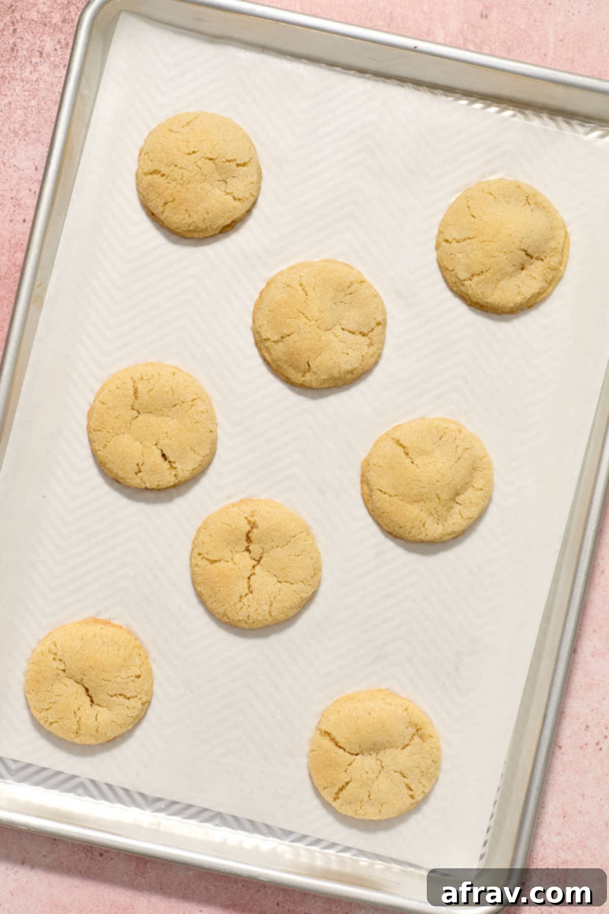 Freshly baked sugar cookies on a baking sheet, showing their puffy shape and crackled tops, with slightly browned edges.