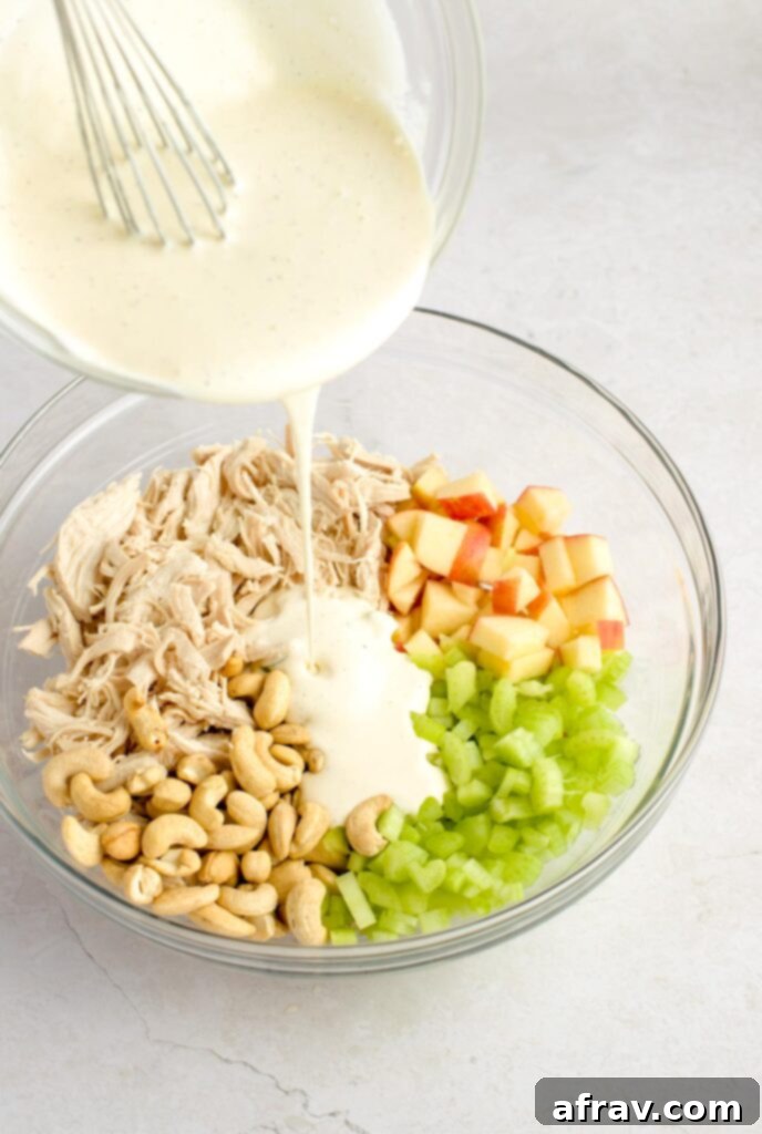 Dairy-free dressing being poured over the chicken, apples, cashews, and celery in a mixing bowl.