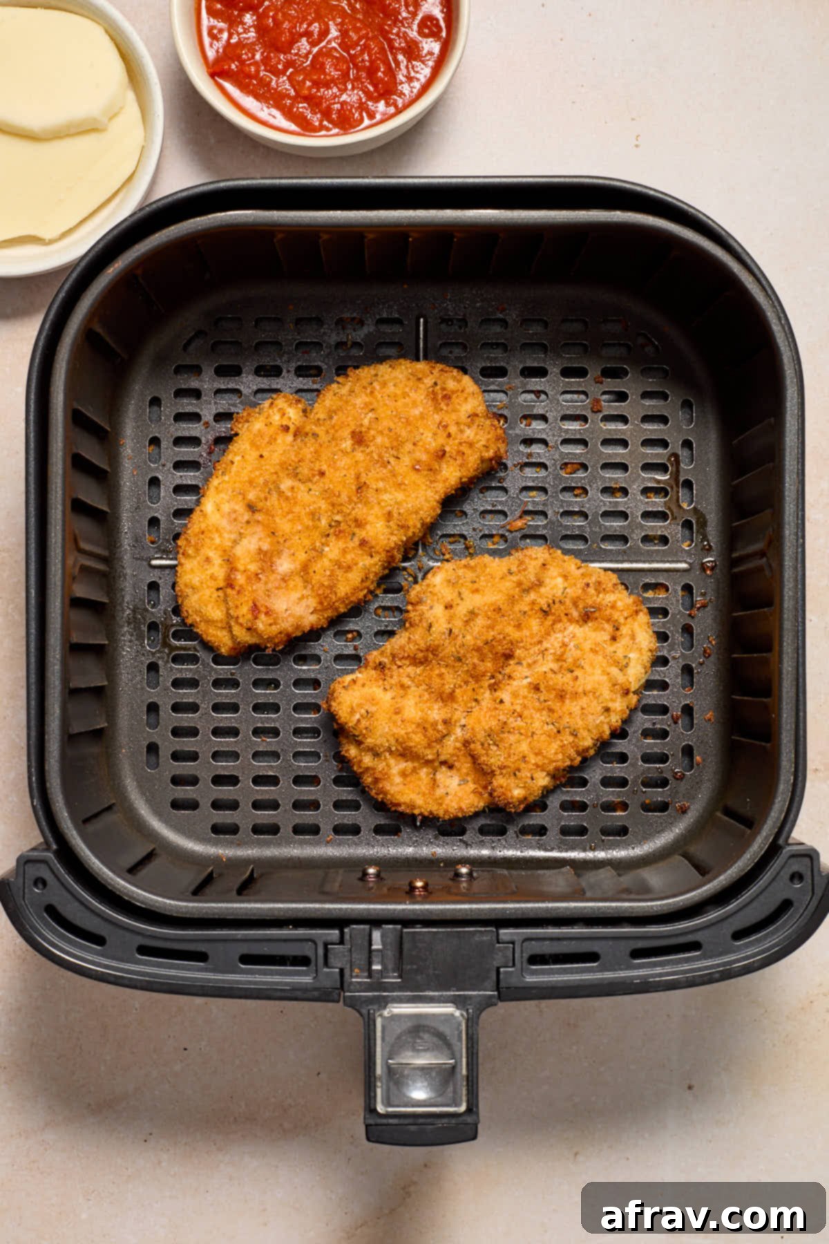 Air fried breaded chicken in the air fryer basket with bowls of marinara and sliced cheese on the side.