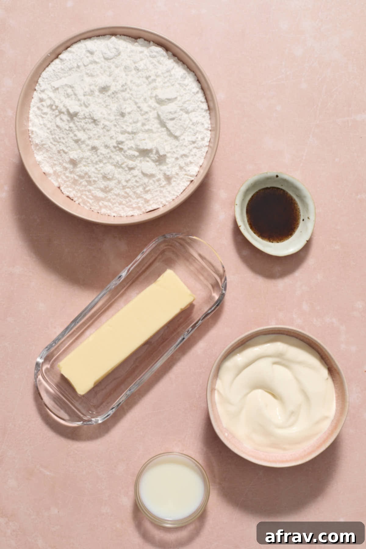 Various ingredients for vanilla sour cream frosting, including butter, sour cream, powdered sugar, vanilla bean paste, and milk, are neatly arranged on a white surface.