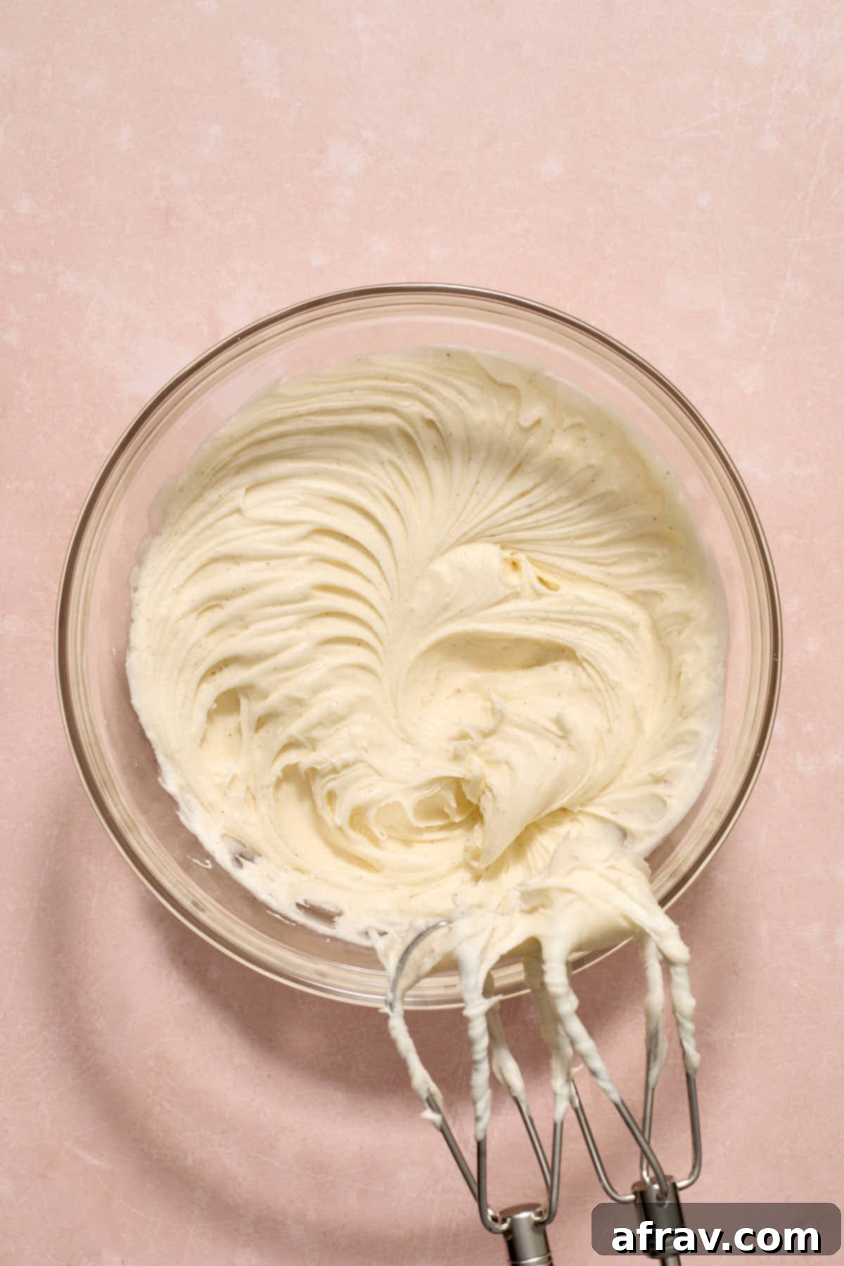 Vanilla bean sour cream frosting actively whipping in a clear glass bowl, showcasing its creamy and fluffy texture.