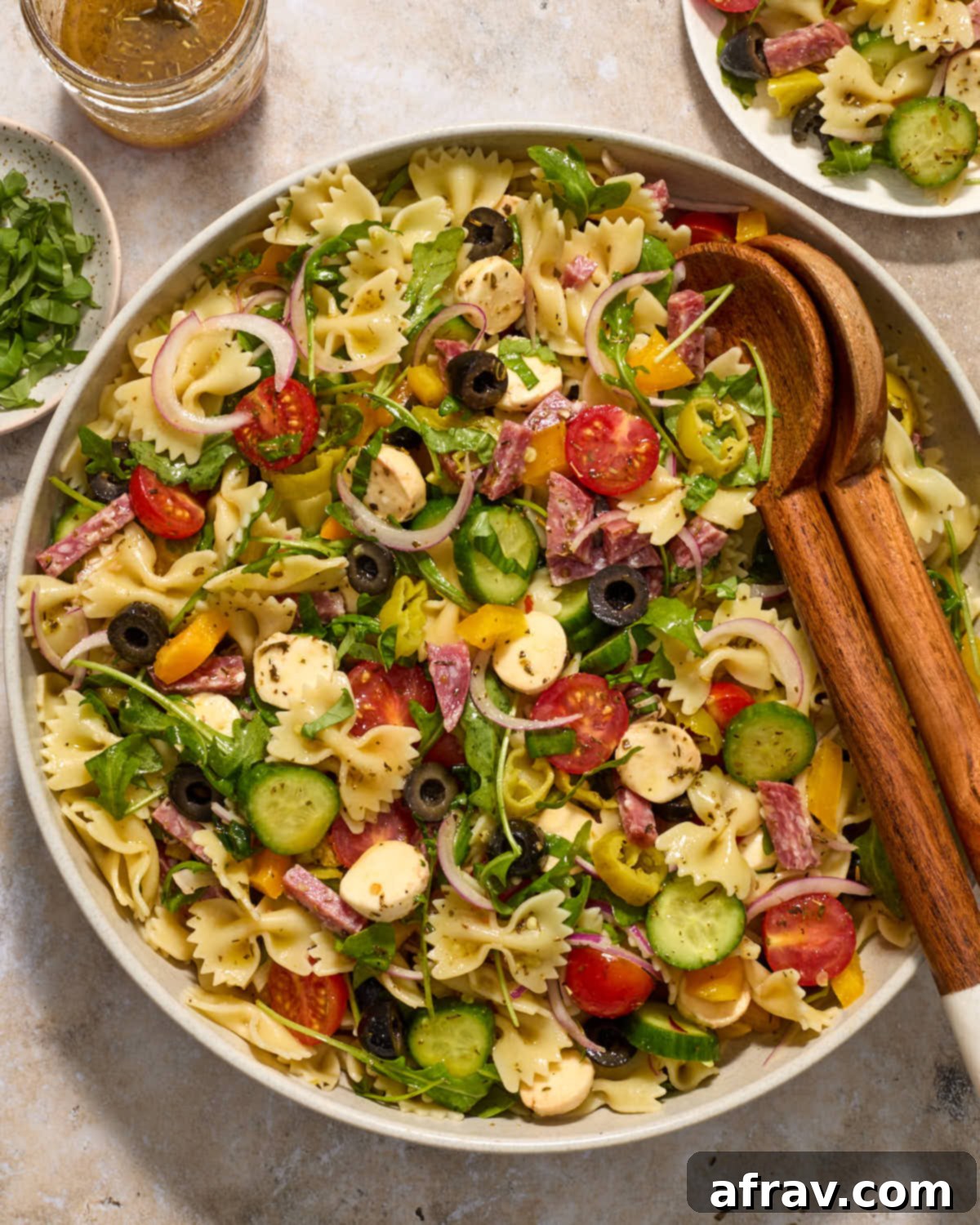 A beautifully presented Italian pasta salad in a bowl, with a plated serving, extra vinaigrette, and fresh basil on the side.