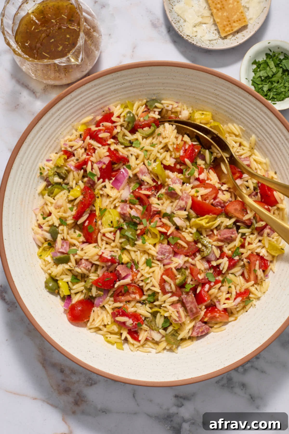 A large bowl of colorful Italian Orzo Salad with a small bowl of dressing, grated parmesan, and chopped parsley on the side, ready to be served.